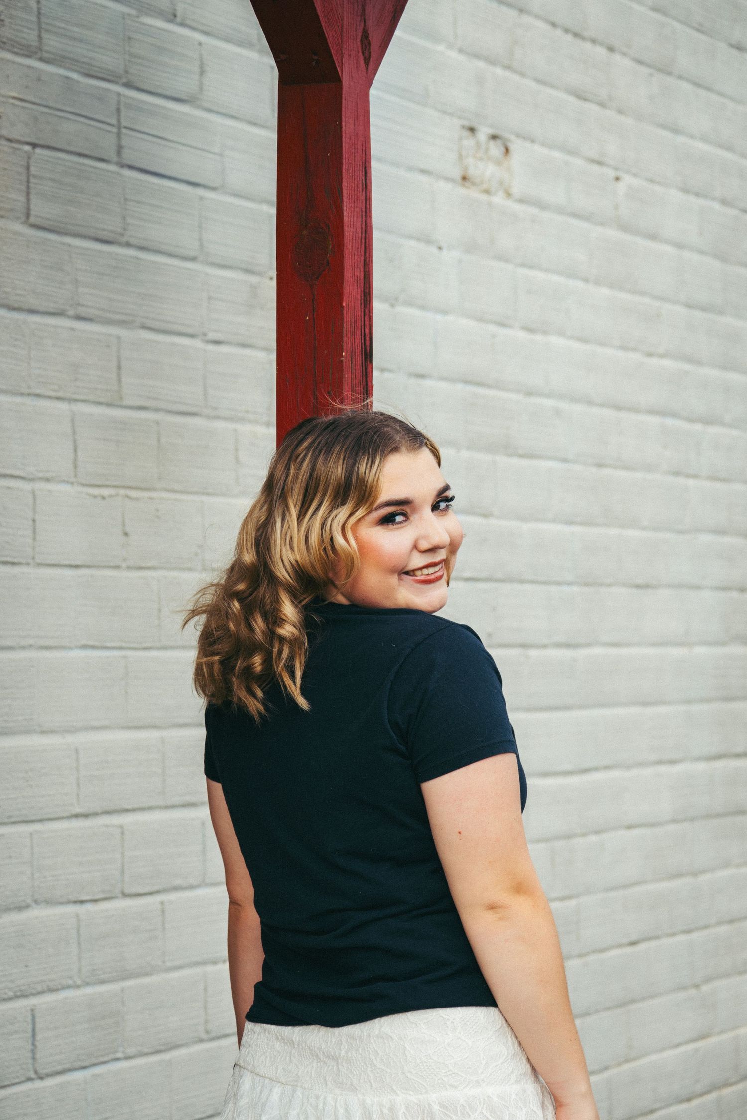 Young woman in black tee and white skirt smiles over shoulder against white brick wall with red beam.