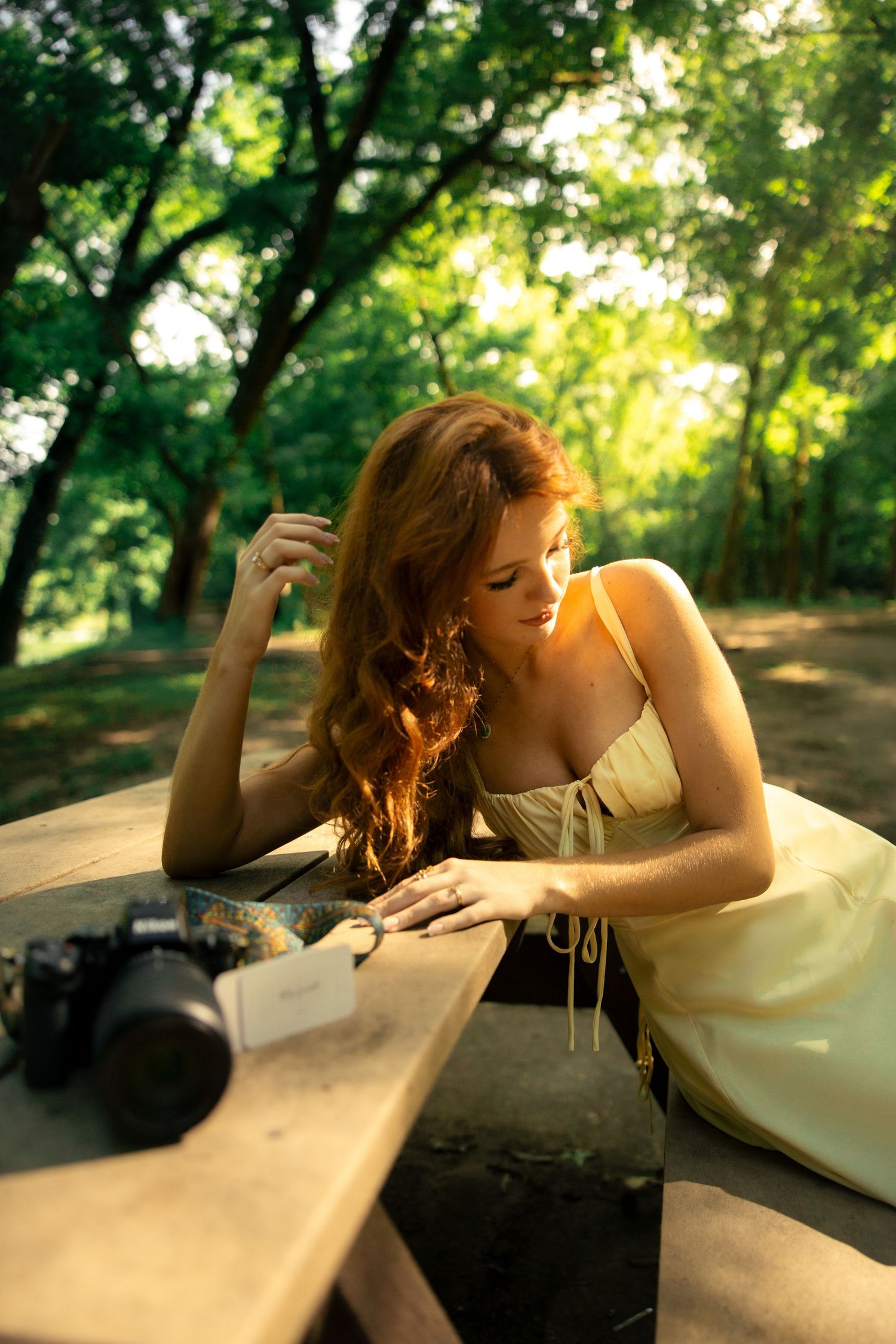 Red-haired woman in yellow dress reading at wooden table in sunlit forest with vintage camera nearby.