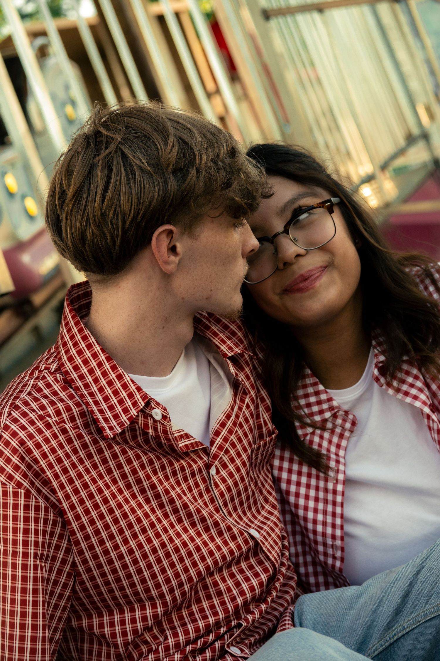 A young couple in matching red plaid shirts share a tender moment together at an amusement park.