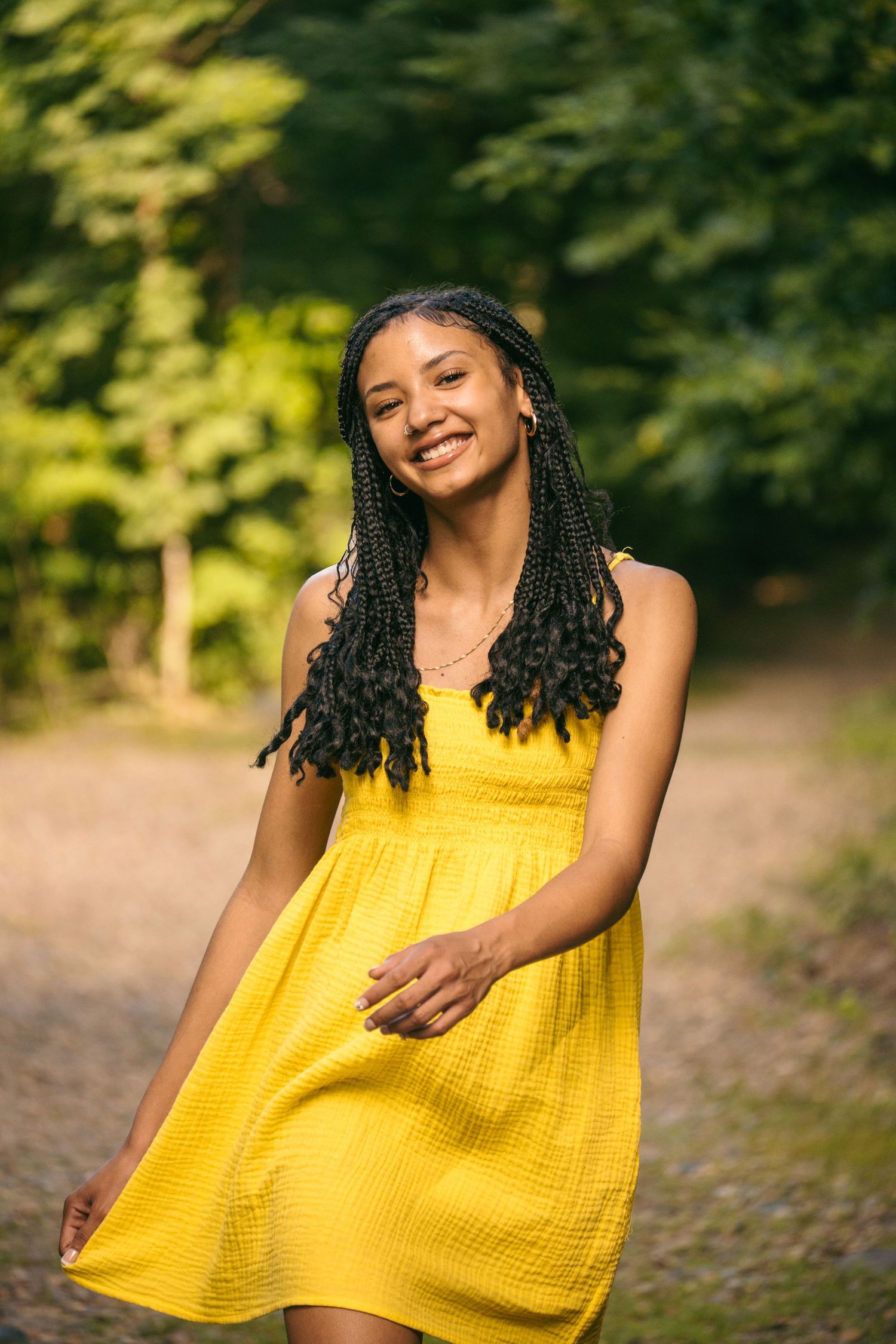 Smiling young woman in a yellow dress walking along a sunlit wooded path.