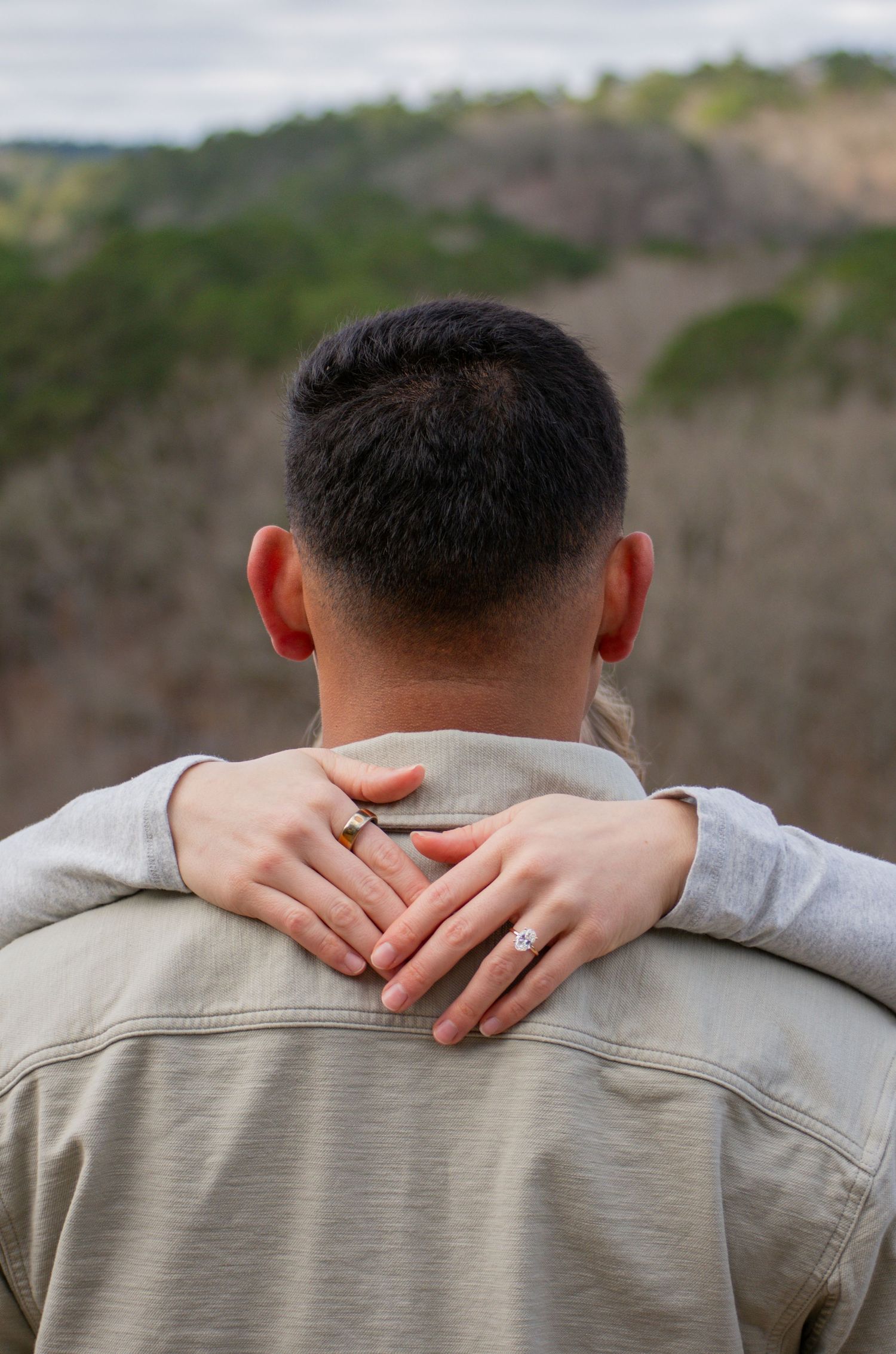 Back view of man with woman's hands showing diamond engagement ring resting on his shoulders.