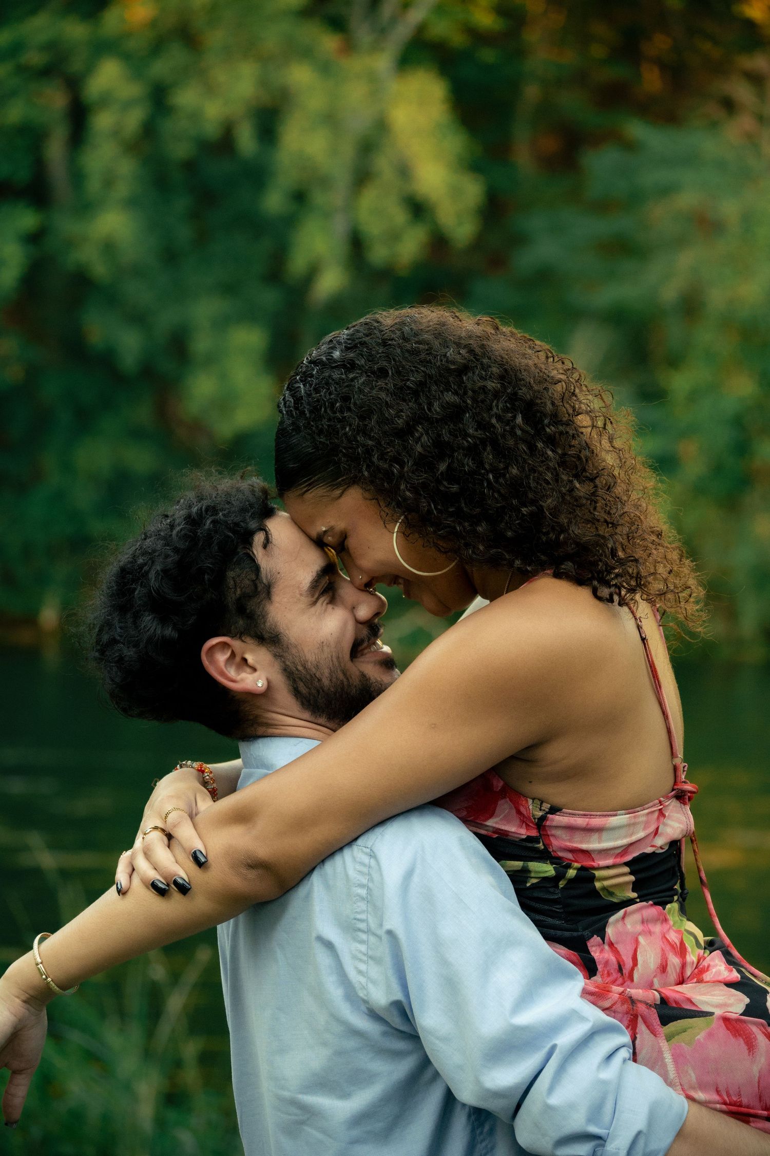 A couple shares a romantic embrace outdoors, with the woman in a floral dress smiling at her partner near a lush green backdrop.