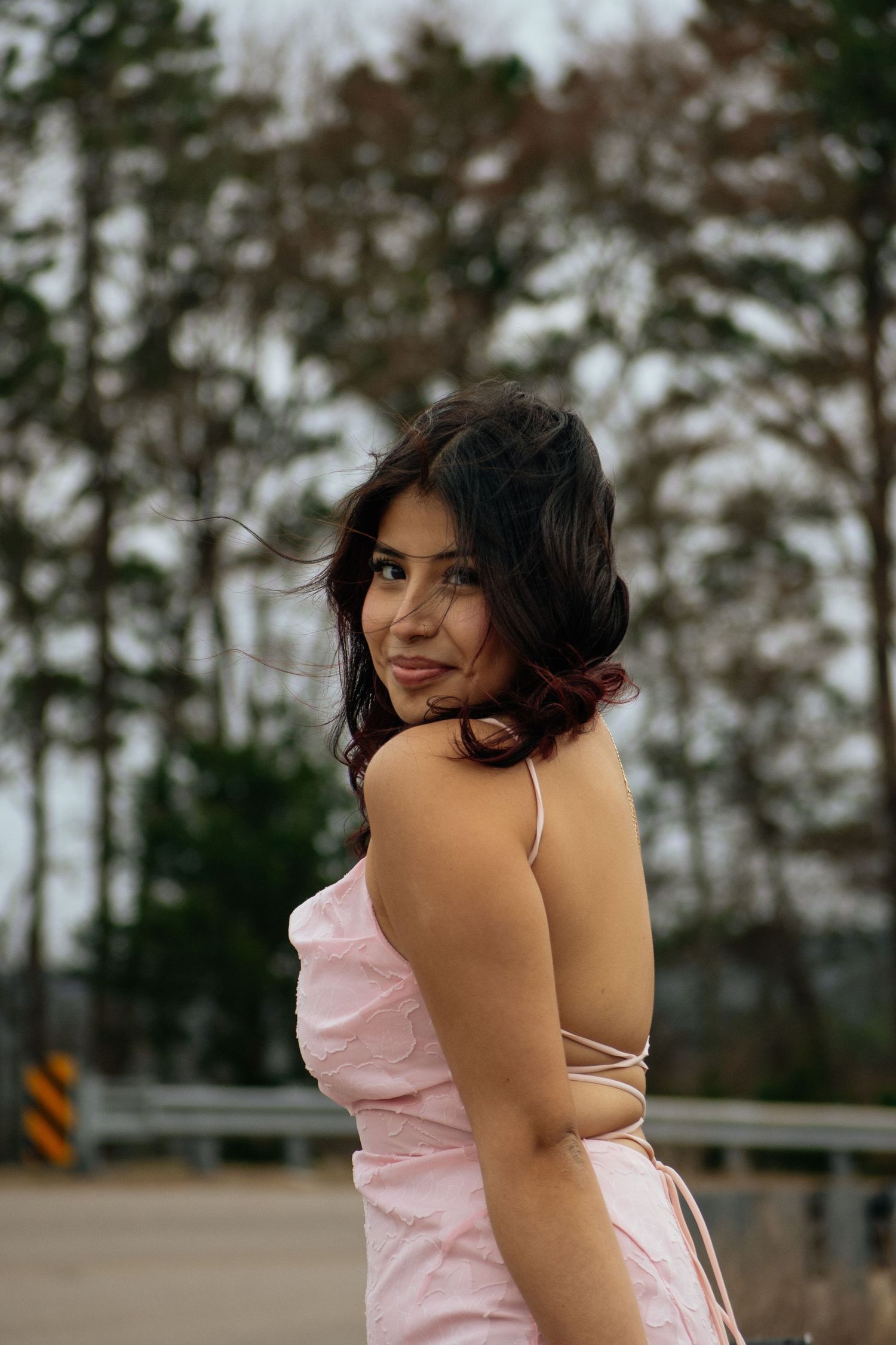 Young woman in pink spaghetti strap dress smiles over shoulder outdoors with pine trees in background.