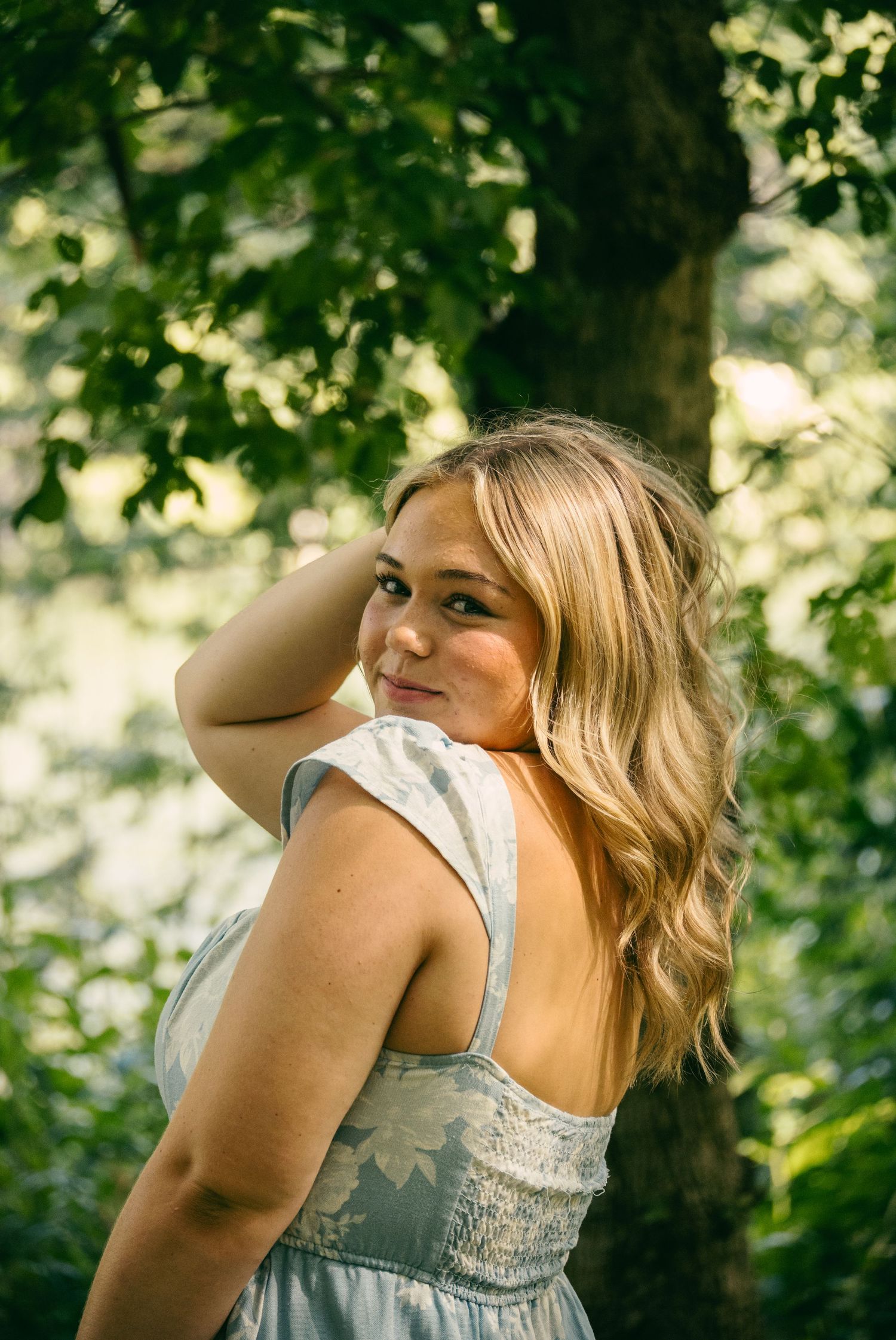 Young blonde woman in a light blue dress posing among green trees in a sunlit forest.