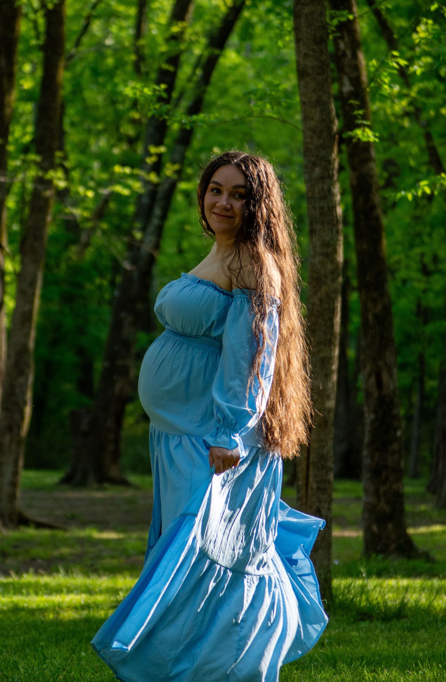Pregnant woman in flowing blue off-shoulder dress twirling in a lush green forest.