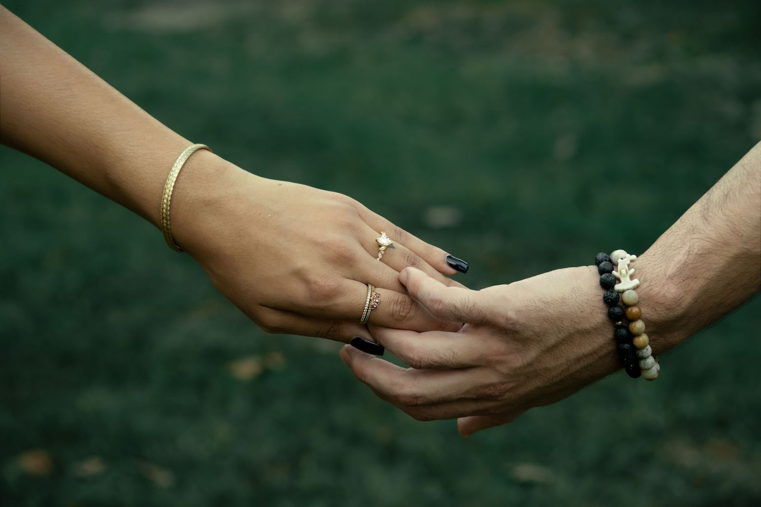 Two hands holding gently, one with gold bracelet and ring, other with beaded bracelet, against green grass background.