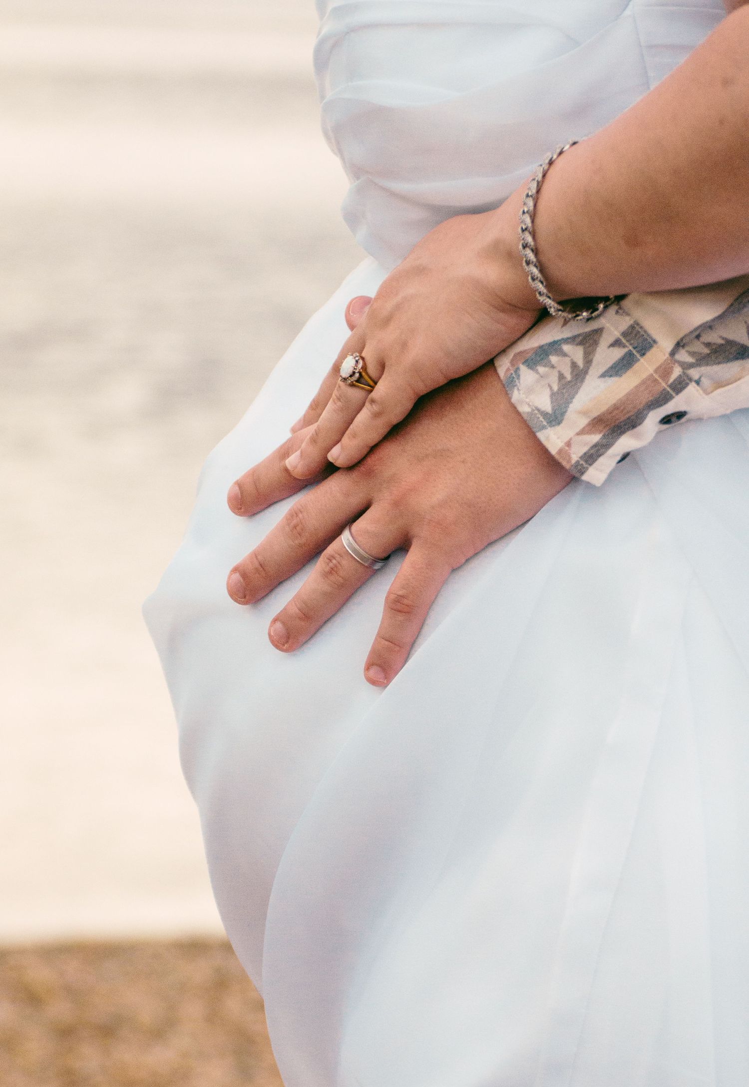 Close-up of bride and groom hands with wedding rings and bracelet on white wedding dress.
