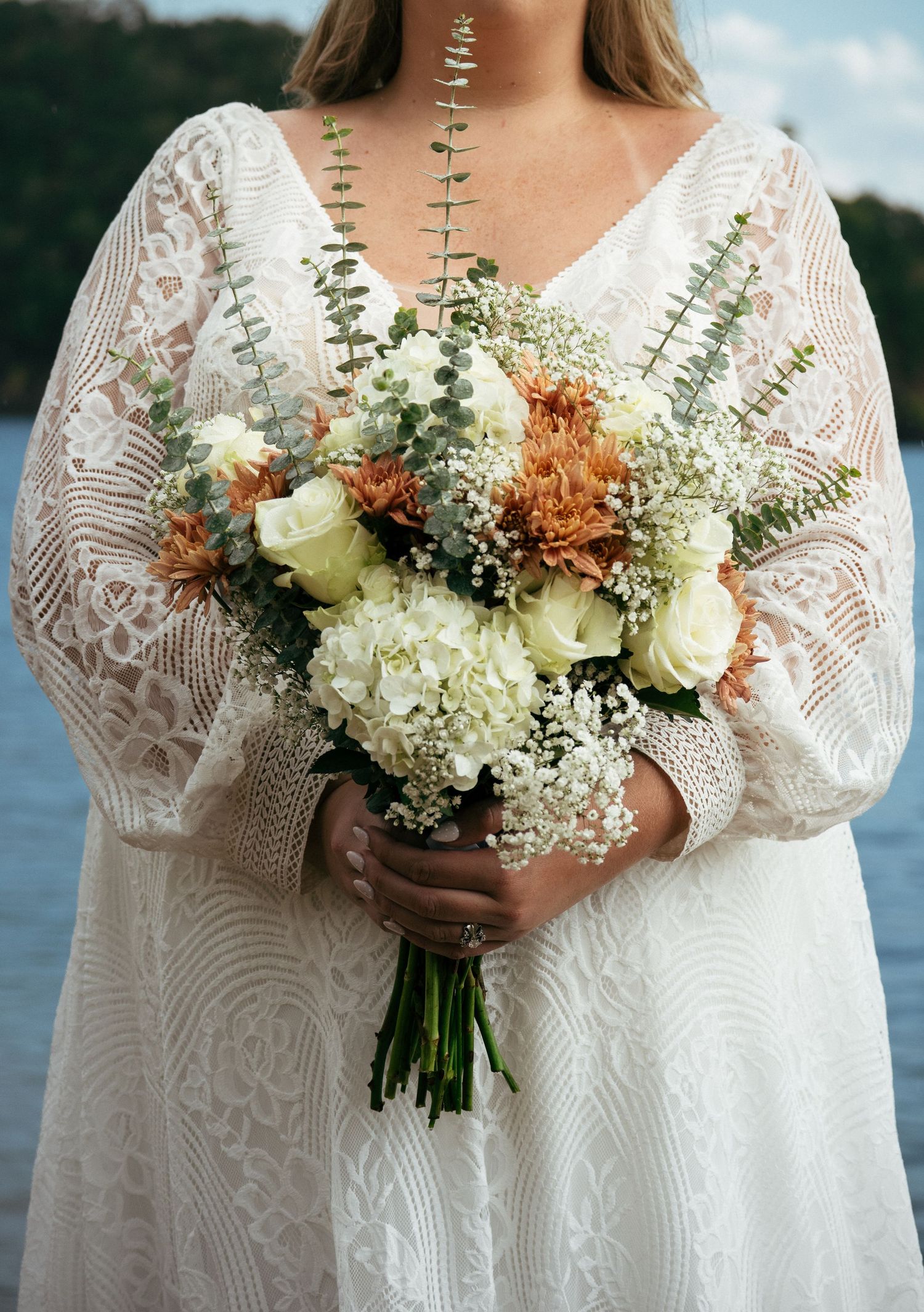 Bride in lace wedding dress holding bouquet of white roses, hydrangeas, orange mums, and eucalyptus by a lake.