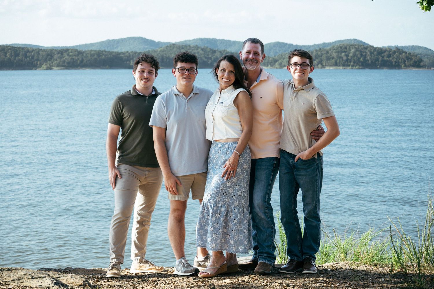 Family of five smiling together on a rocky lakeside shore with scenic blue water and green hills in the background.