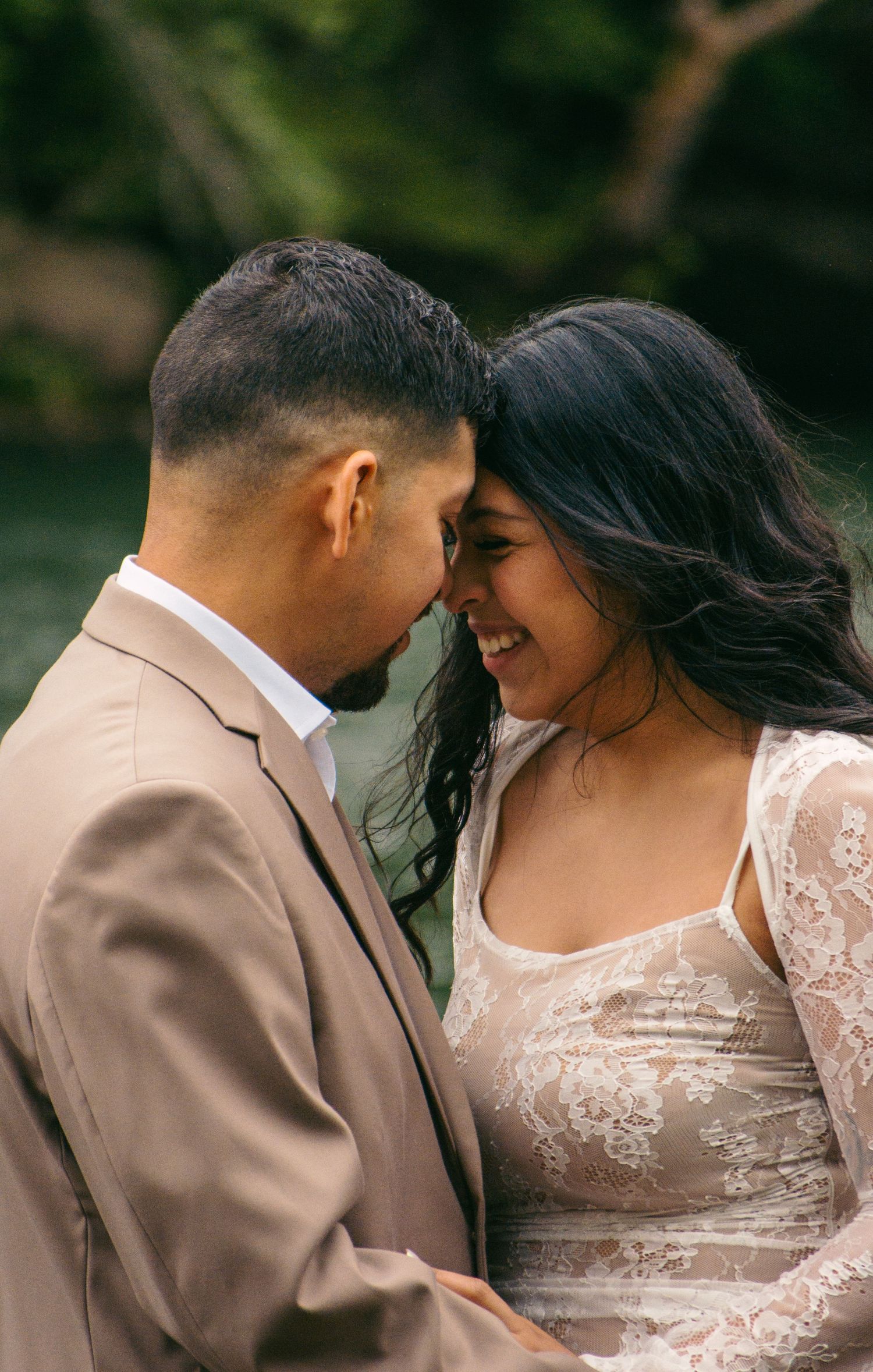 Bride and groom in lace wedding dress and tan suit sharing an intimate forehead touch outdoors.