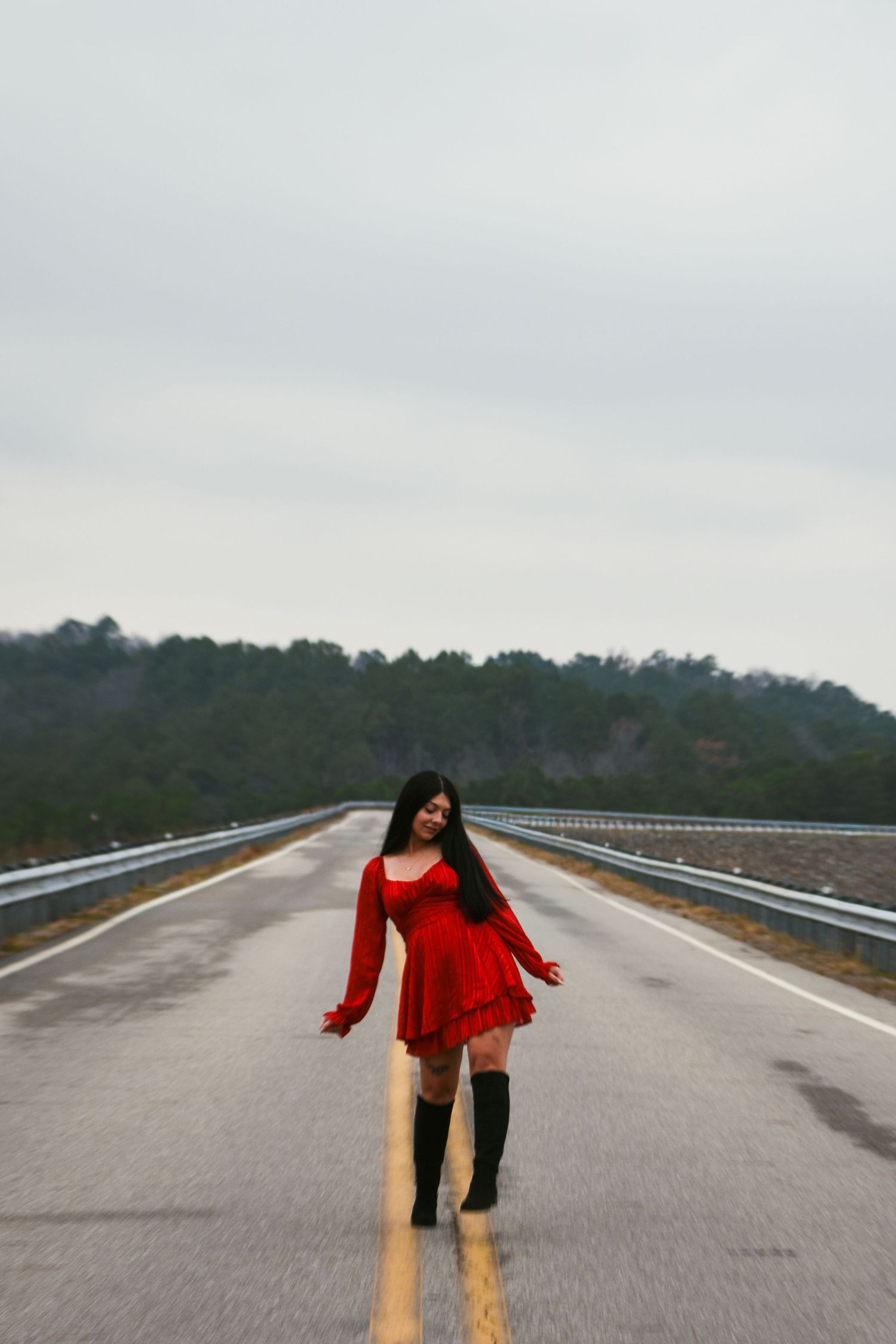 Woman in red mini dress and black boots standing on empty road with overcast sky and trees.