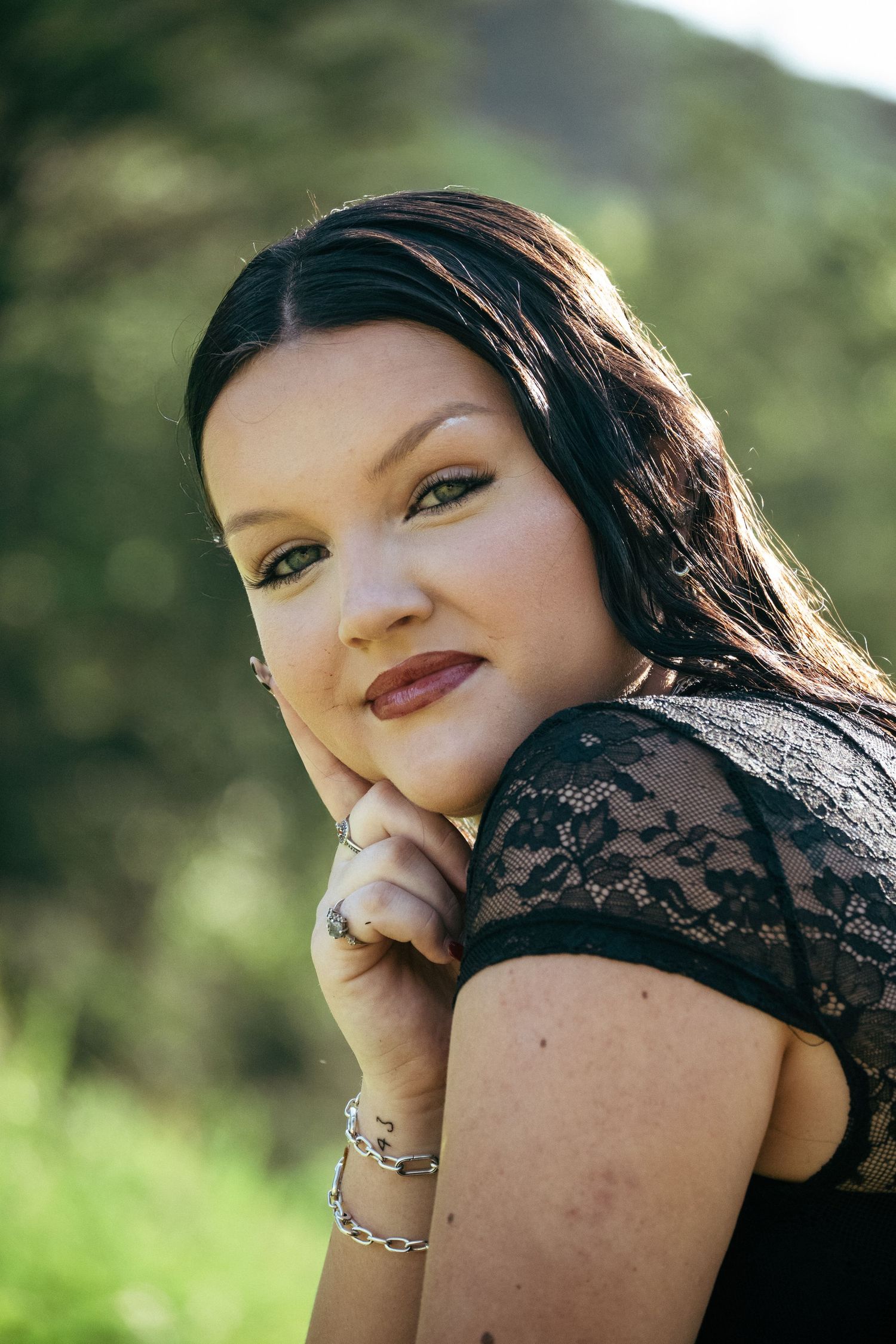 Young woman with dark hair in black lace top poses outdoors with hand on chin, wearing silver bracelet.
