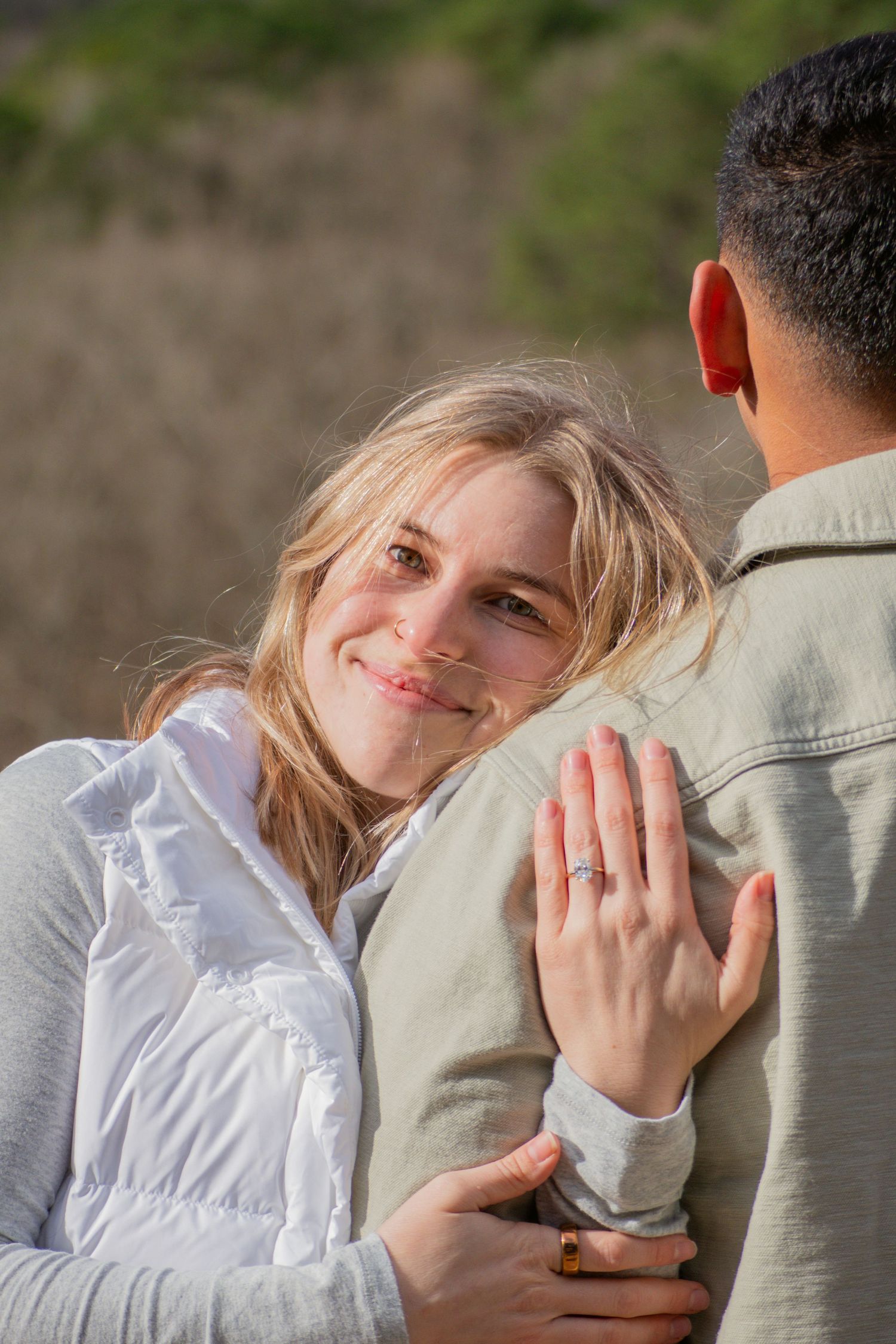 Smiling blonde woman showing engagement ring while embracing partner outdoors in natural setting.