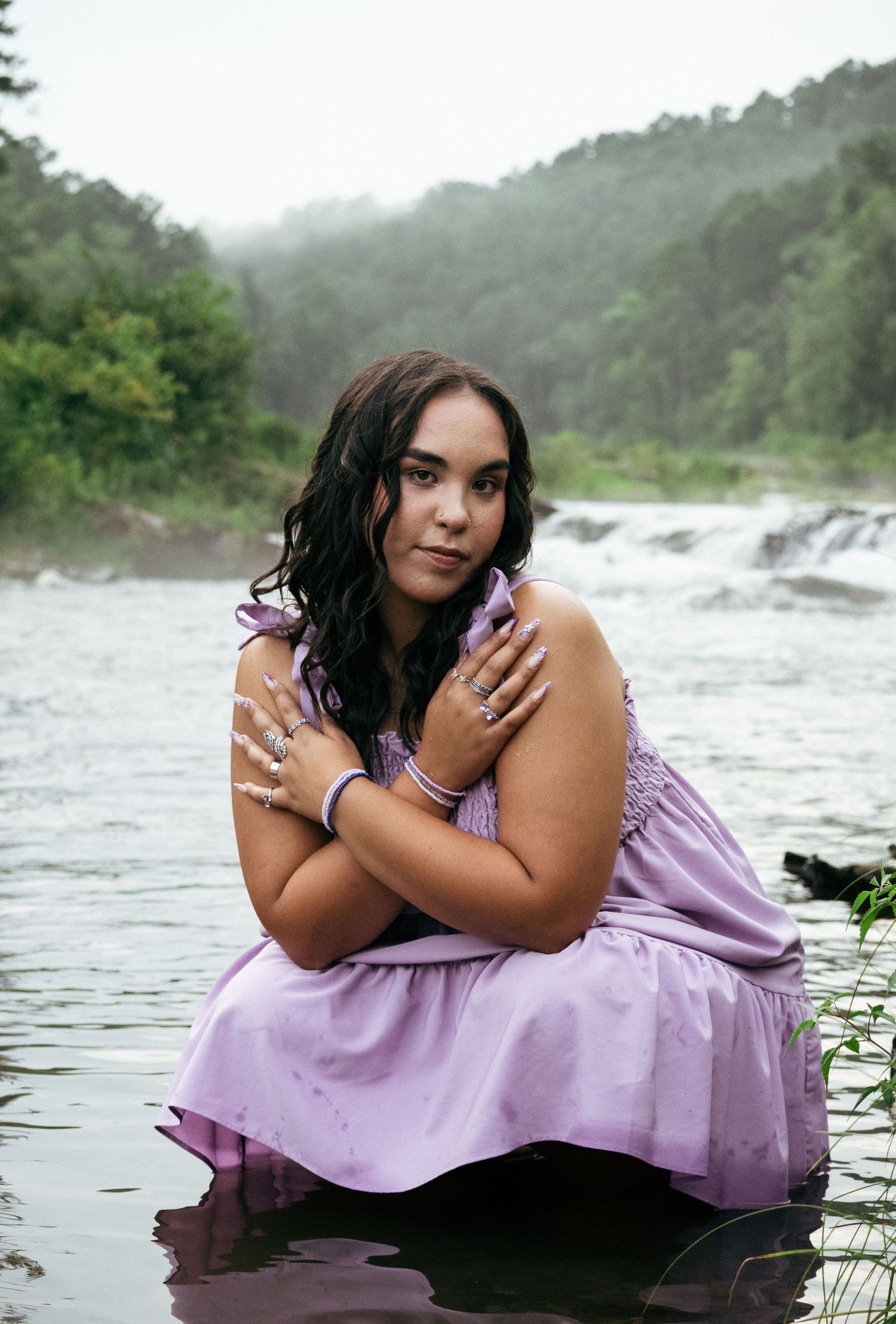 Young woman in lavender dress sitting in a river with a waterfall and lush green forest in the background.