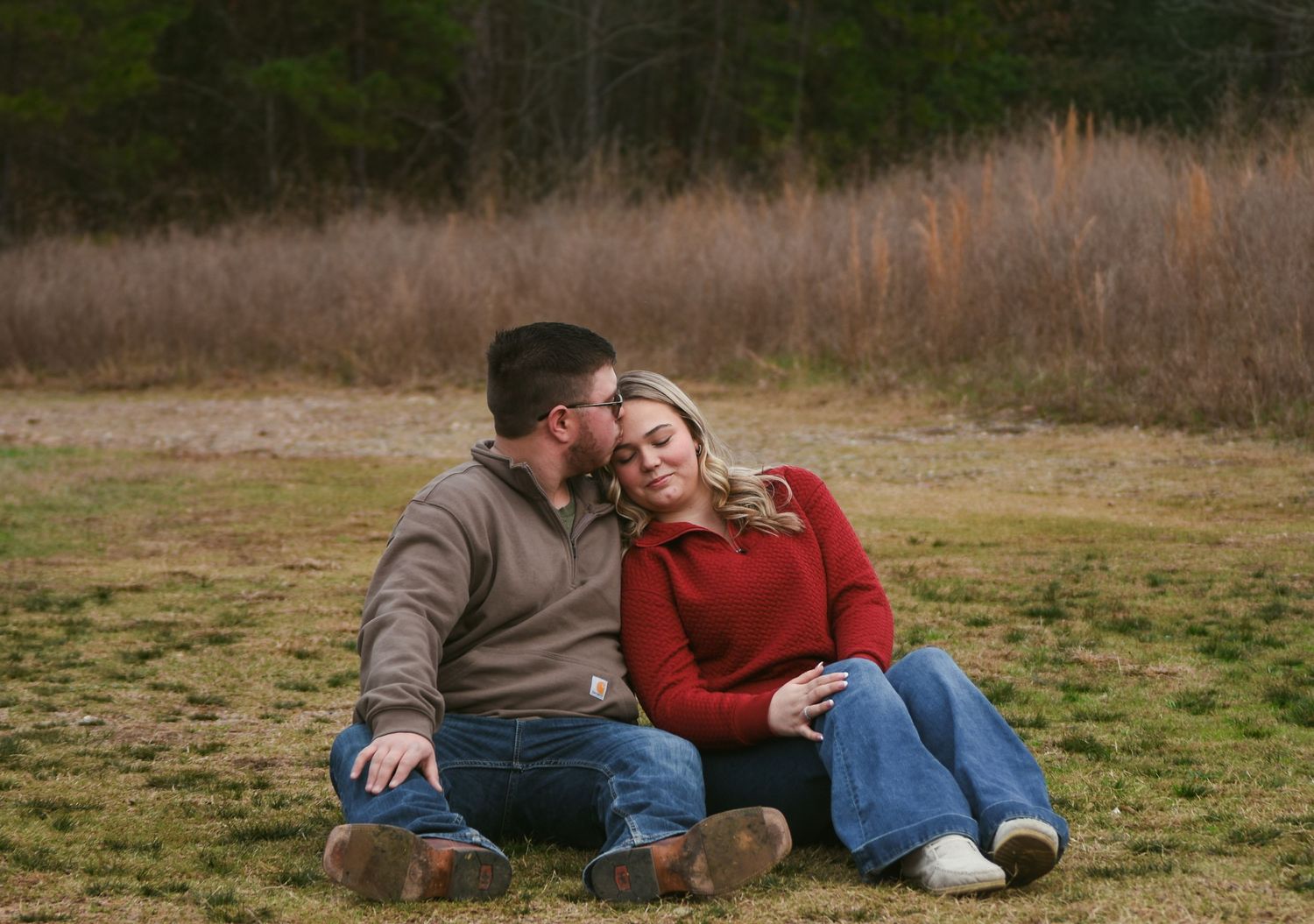 Romantic couple sitting on grass, man kissing woman on the head, both in casual fall outdoor attire.