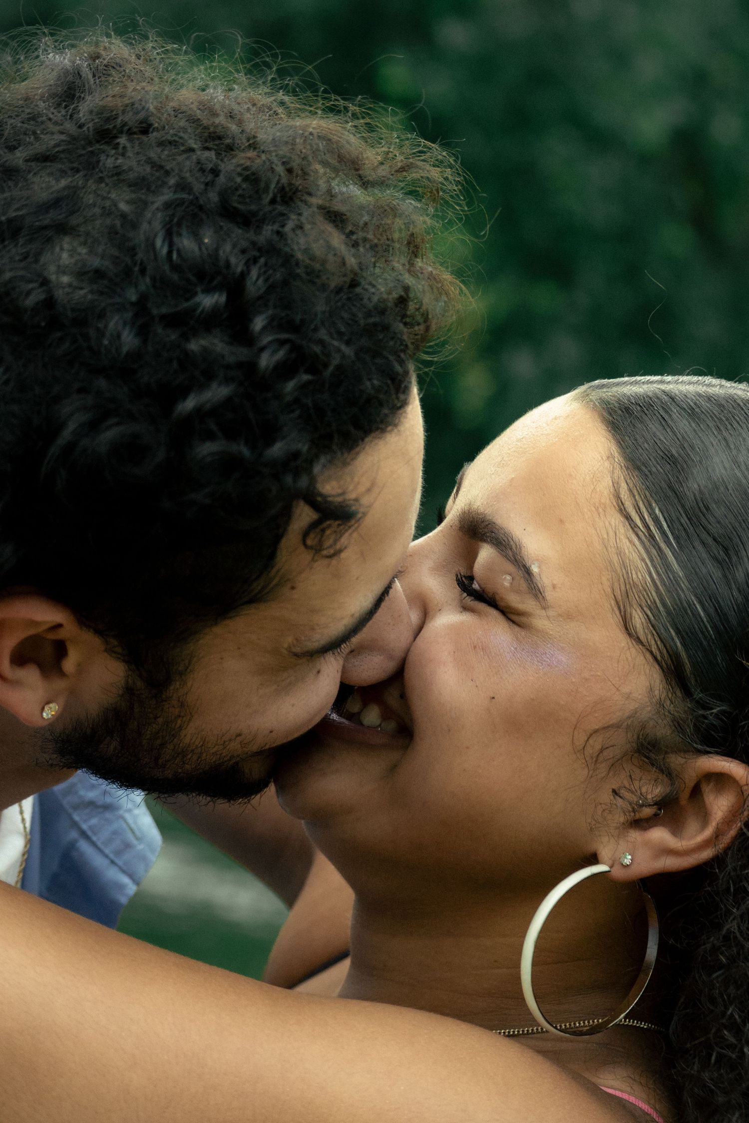 A couple sharing a tender kiss outdoors, with the woman wearing gold hoop earrings and the man having curly hair.