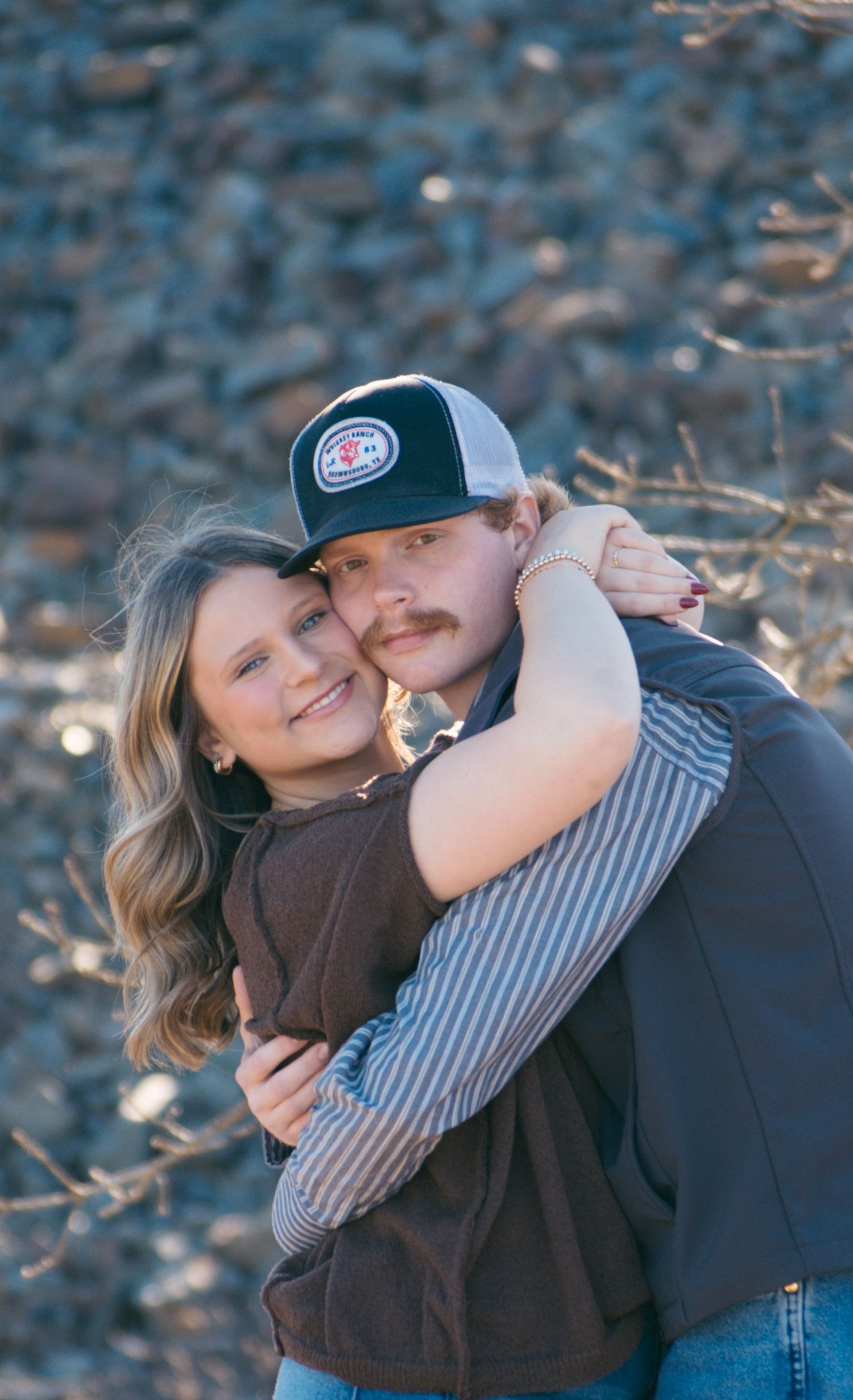 Smiling couple embracing outdoors with woman hugging man in trucker hat against rocky terrain.