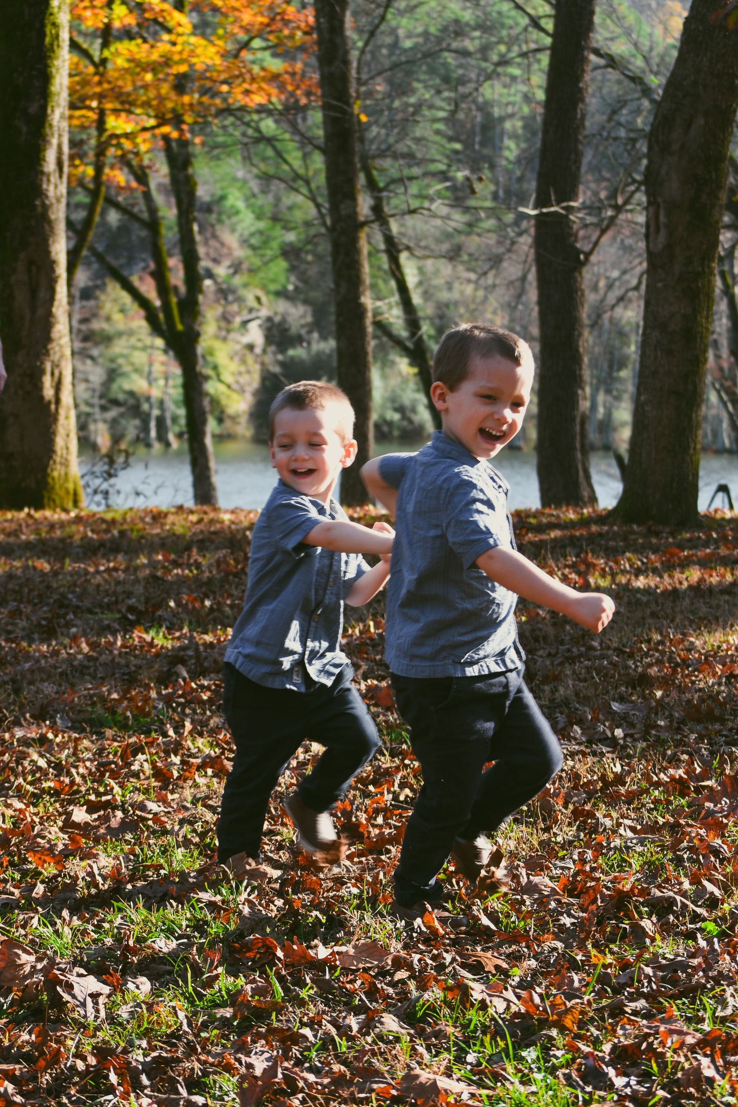 Two laughing children in matching blue shirts running through autumn leaves in a sunlit park.
