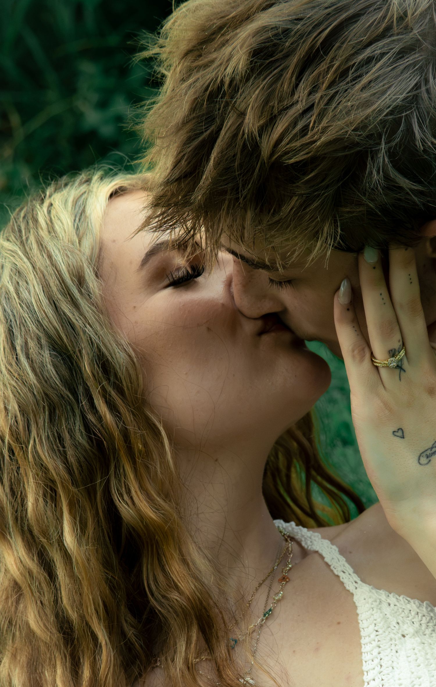 A young couple shares a romantic kiss outdoors, surrounded by lush green foliage, with visible tattoos and jewelry.