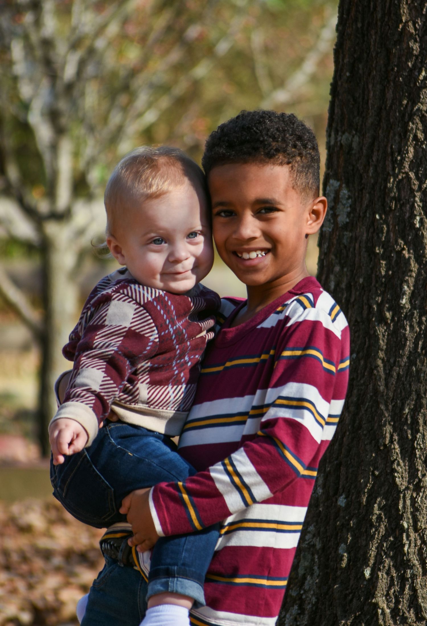 Older boy in striped shirt holding smiling toddler while leaning against a tree in autumn park.