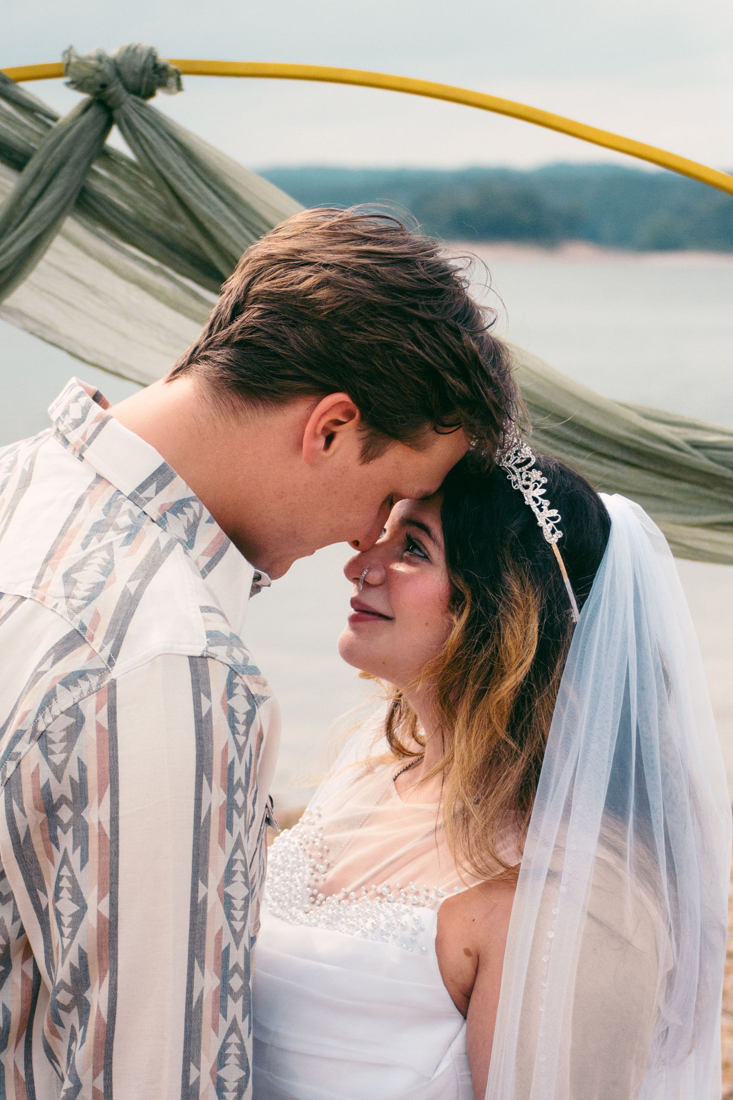 Bride in tiara and veil gazing up at groom under draped fabric arch near waterfront.