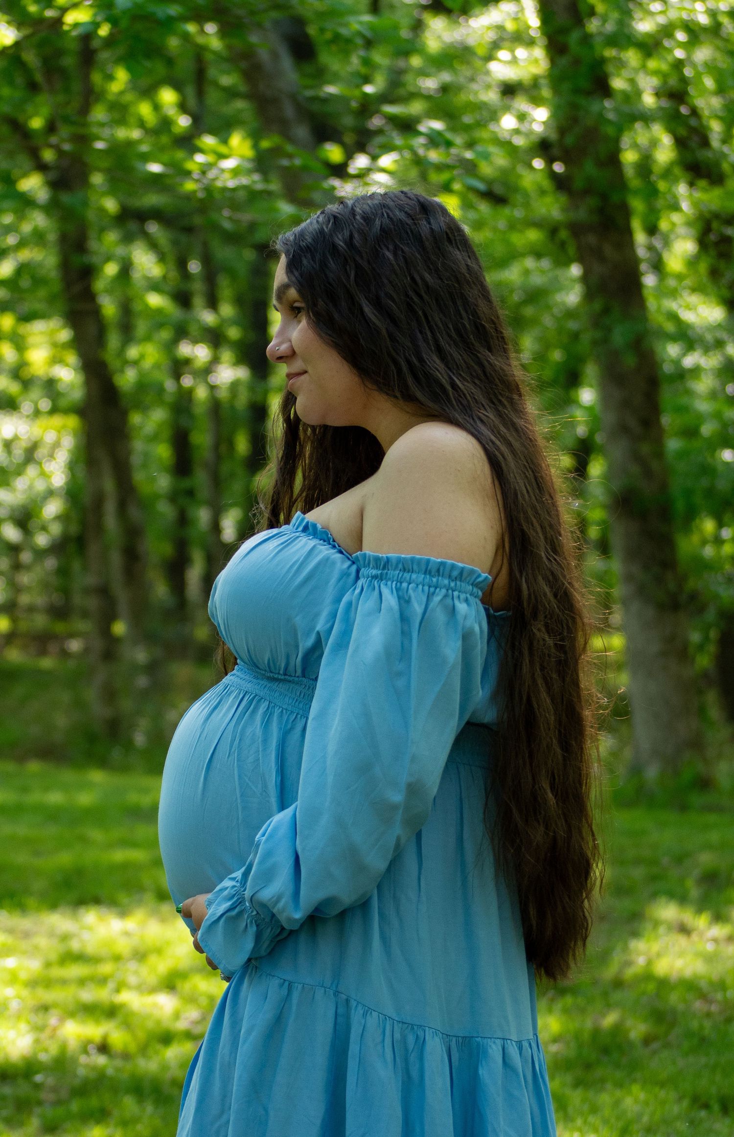 Pregnant woman in blue off-shoulder dress cradling belly while standing sideways in green woods.