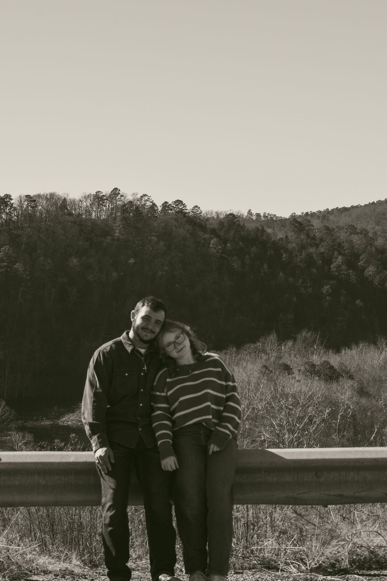 Couple leaning against a guardrail in a black and white photo with forested hills behind them.