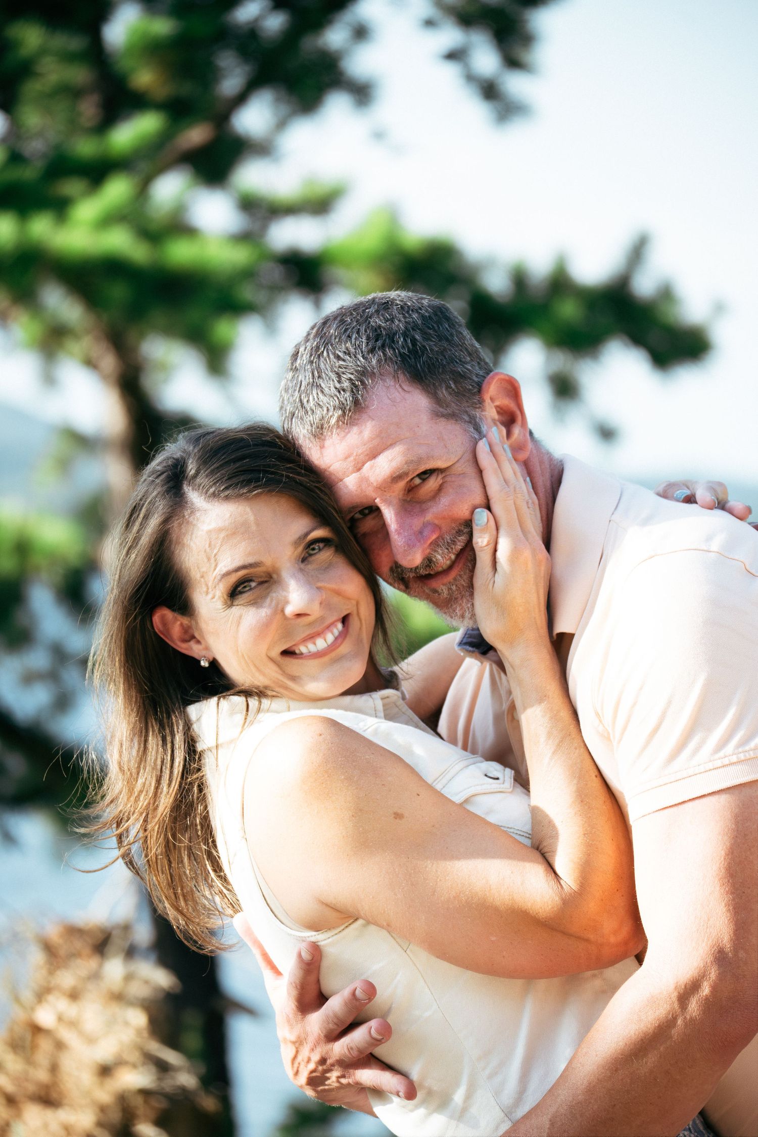 Happy couple embracing outdoors, woman smiling at camera, man hugging her from behind near green trees.