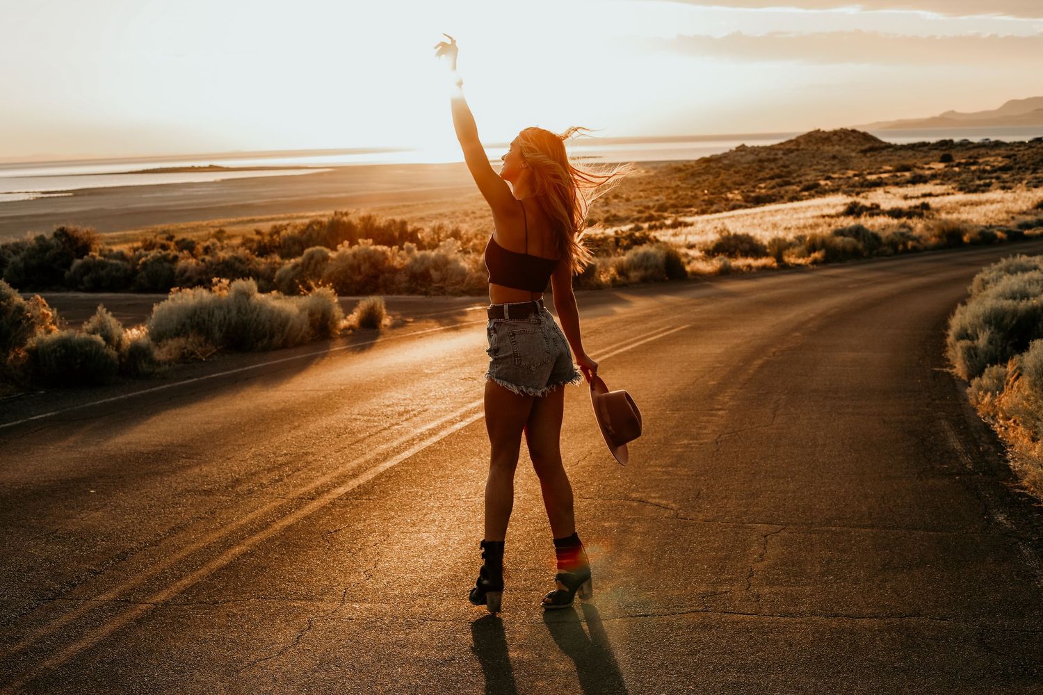 Woman with raised arm standing on open desert road at golden sunset holding a camera.