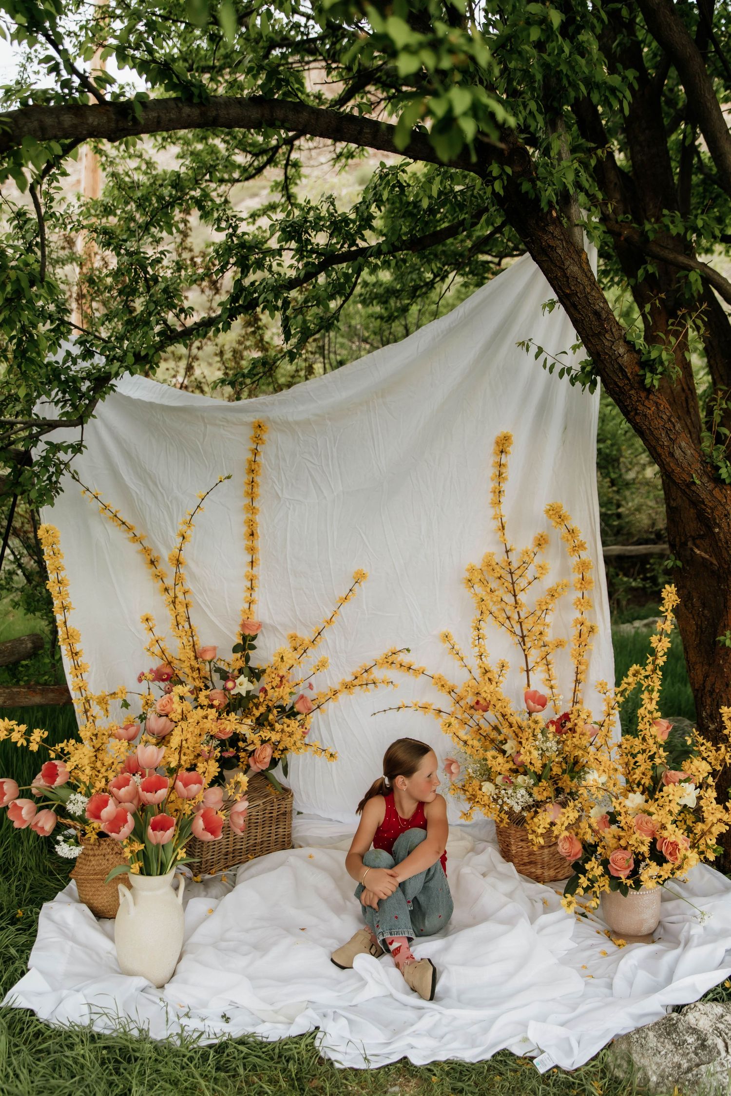 Woman sitting among yellow and pink floral arrangements on white blanket outdoors.