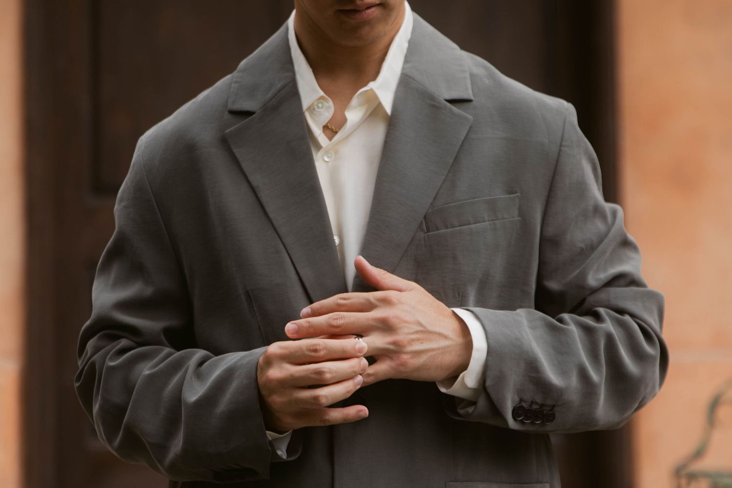 Man in gray suit jacket clasping hands, wearing white dress shirt, standing outdoors near brick wall.
