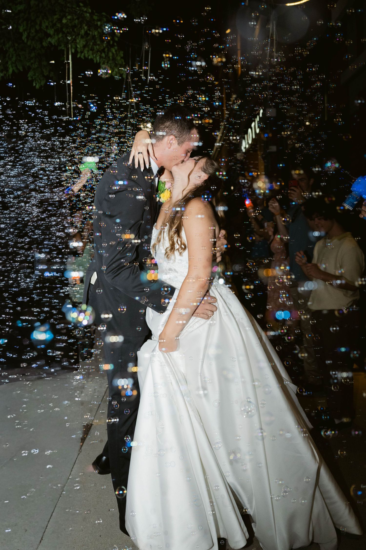 Bride and groom share a romantic kiss surrounded by floating bubbles during their outdoor wedding night celebration.