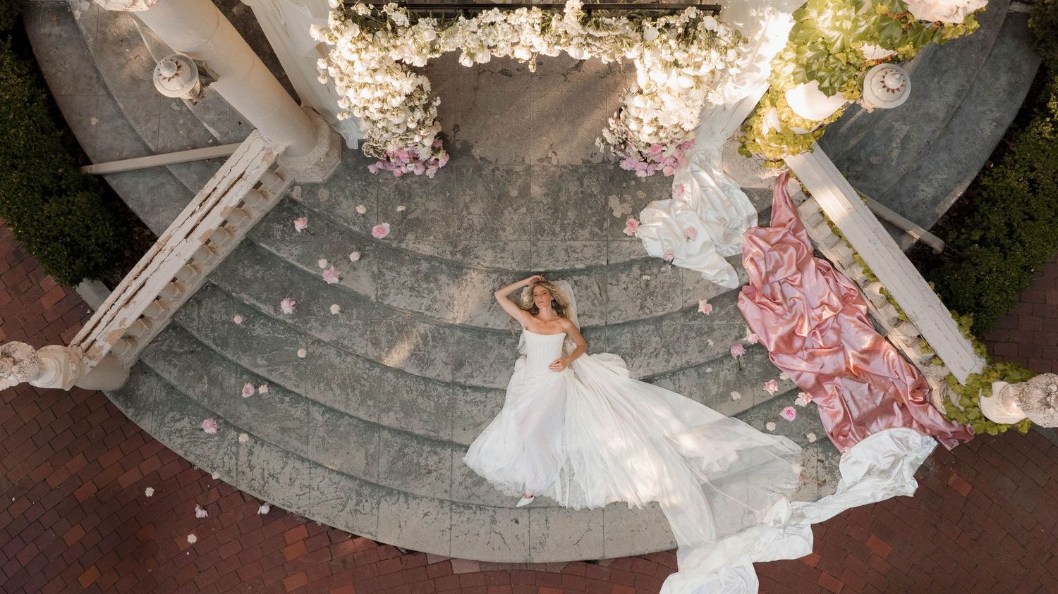 Aerial view of bride in white gown on ornate stone steps adorned with pink floral decorations.