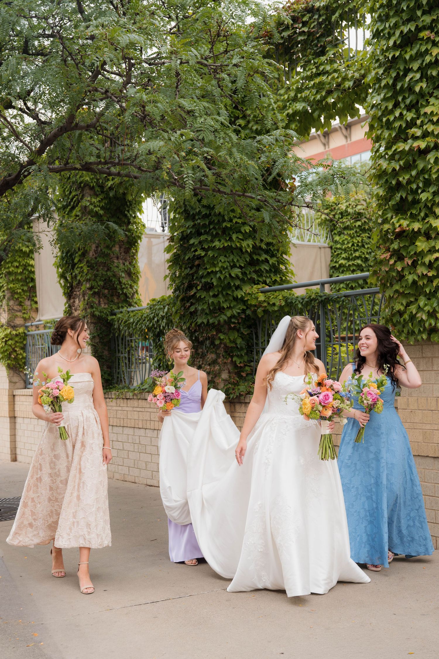 Bride in white gown walks with three bridesmaids in colorful dresses, holding vibrant bouquets outdoors.