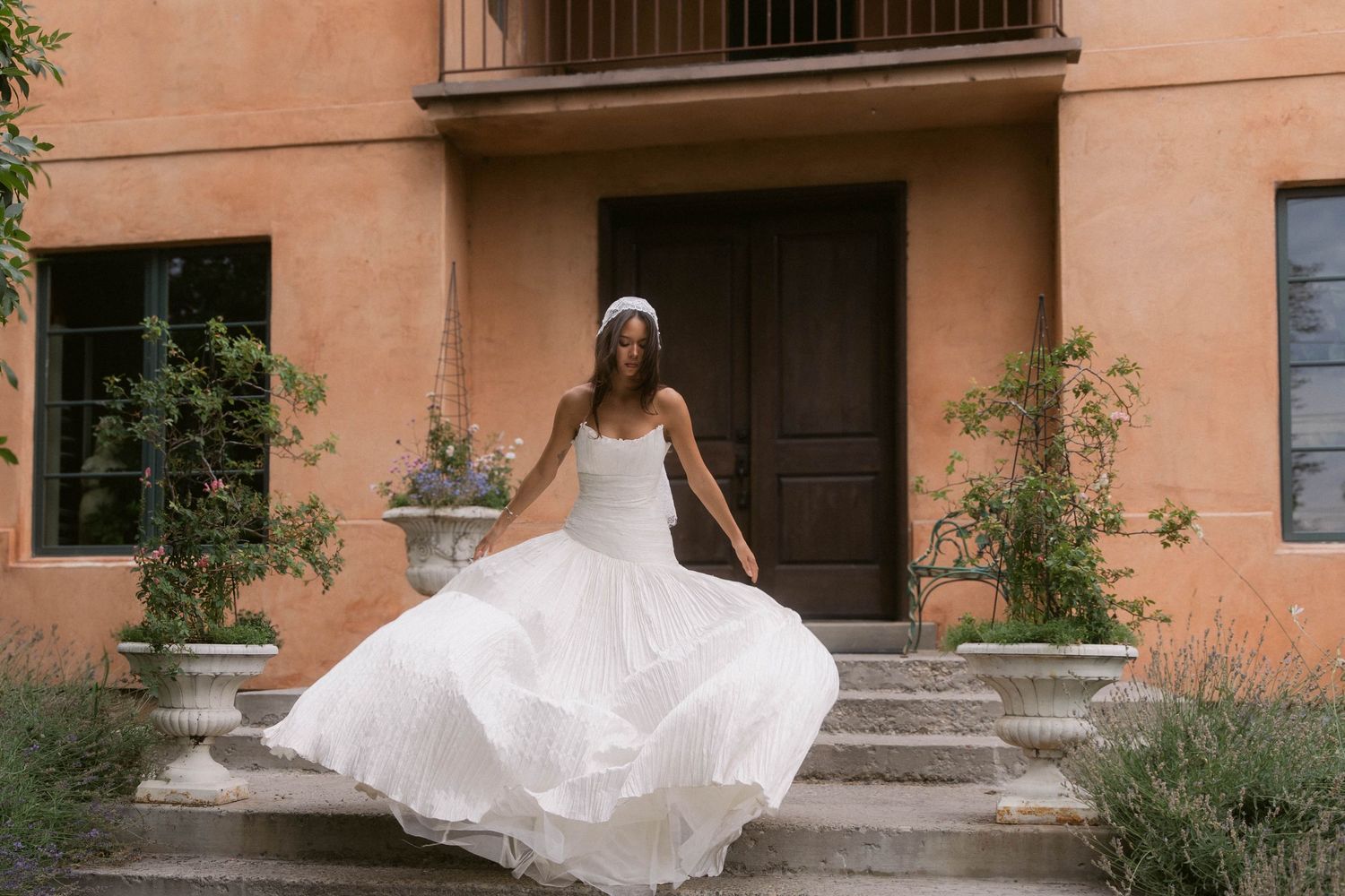 Bride in a flowing white wedding gown twirling on steps of a rustic terracotta villa surrounded by lush greenery.