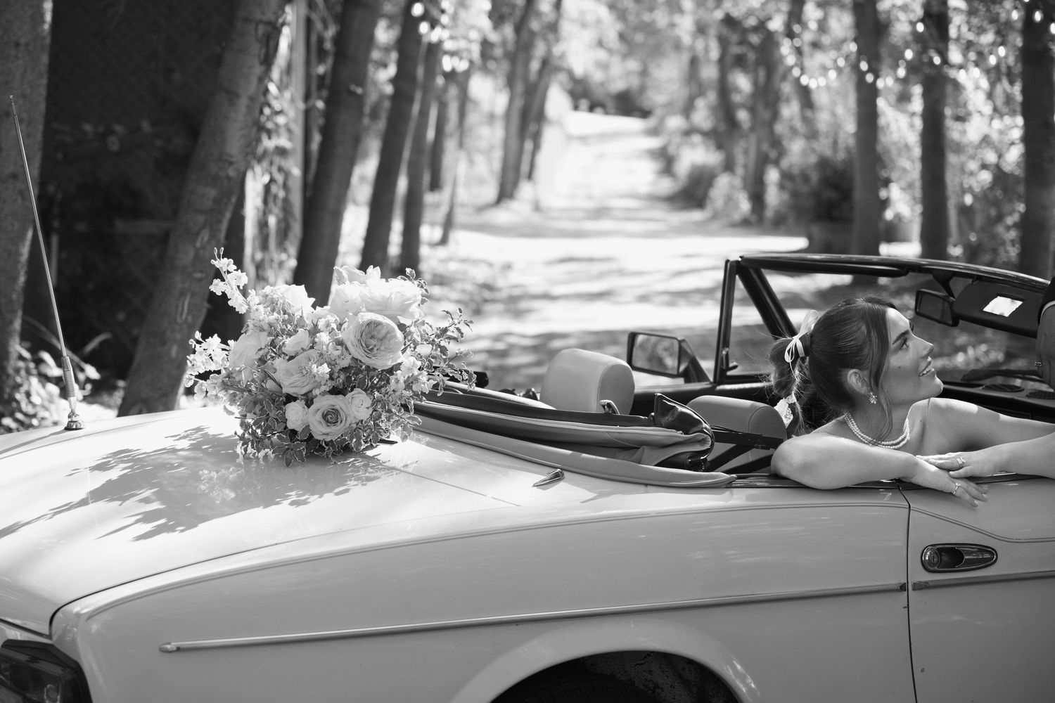 Black and white photo of a bridal bouquet on a vintage convertible car on a tree-lined road.