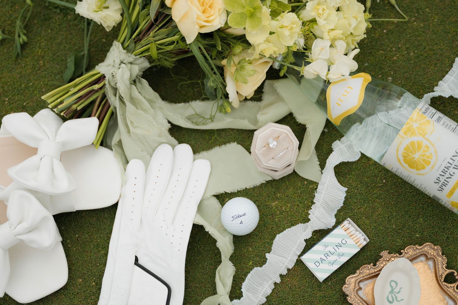 Wedding flat lay featuring white golf gloves, yellow bouquet, golf ball, and sage ribbon on green turf.