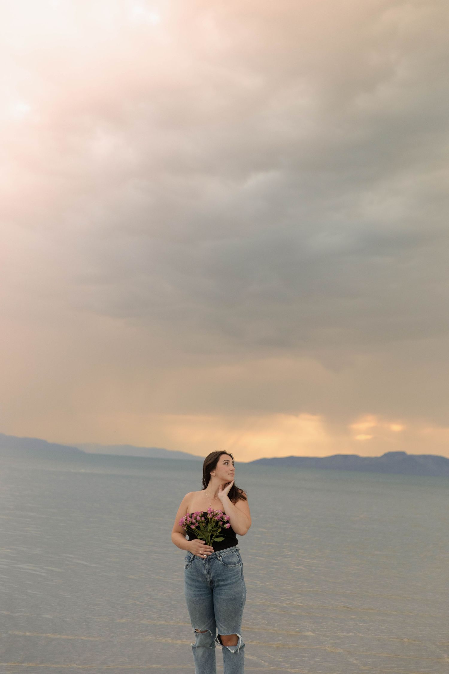 Young woman in ripped jeans holding pink flowers stands by calm lake under dramatic cloudy sunset sky.