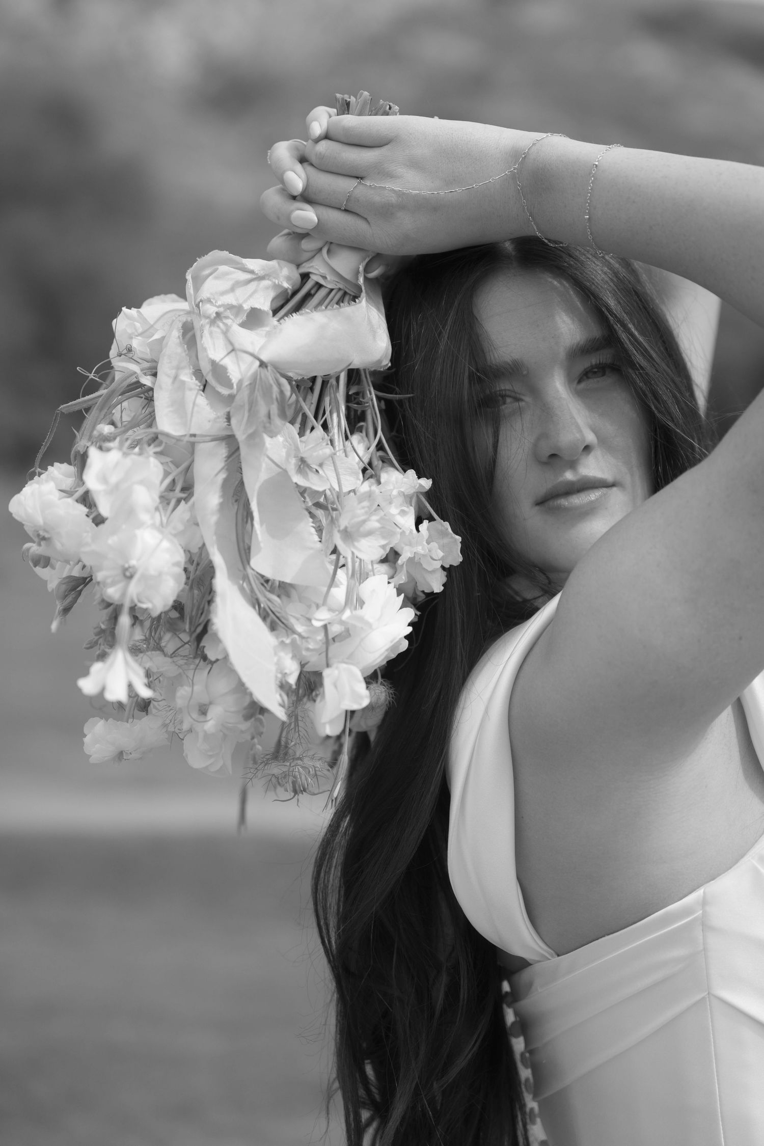 Black and white portrait of dark-haired bride holding white floral bouquet above her head outdoors.