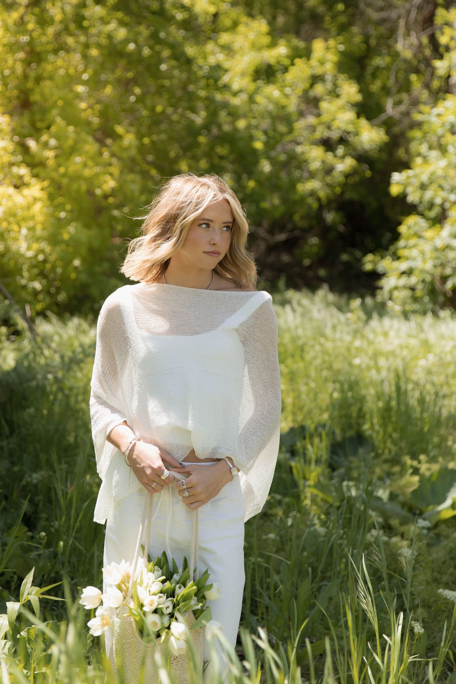 Blonde bride in white dress and knit wrap holds white tulip bouquet in lush green outdoor setting.