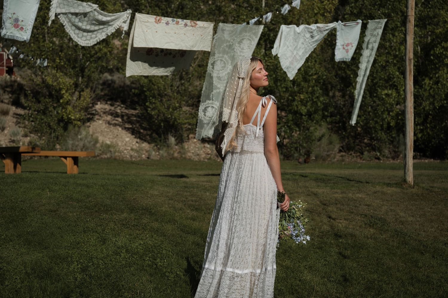 Bride in backless lace wedding gown holding bouquet looking up at hanging vintage linens at outdoor wedding.