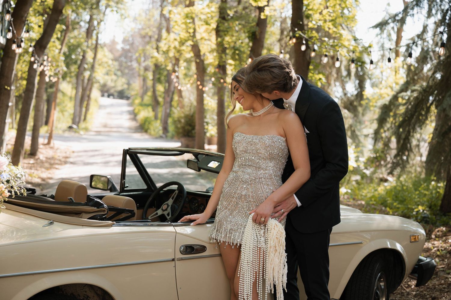 Bride in a beaded fringe dress and groom embrace beside a vintage convertible in a sunlit forest.