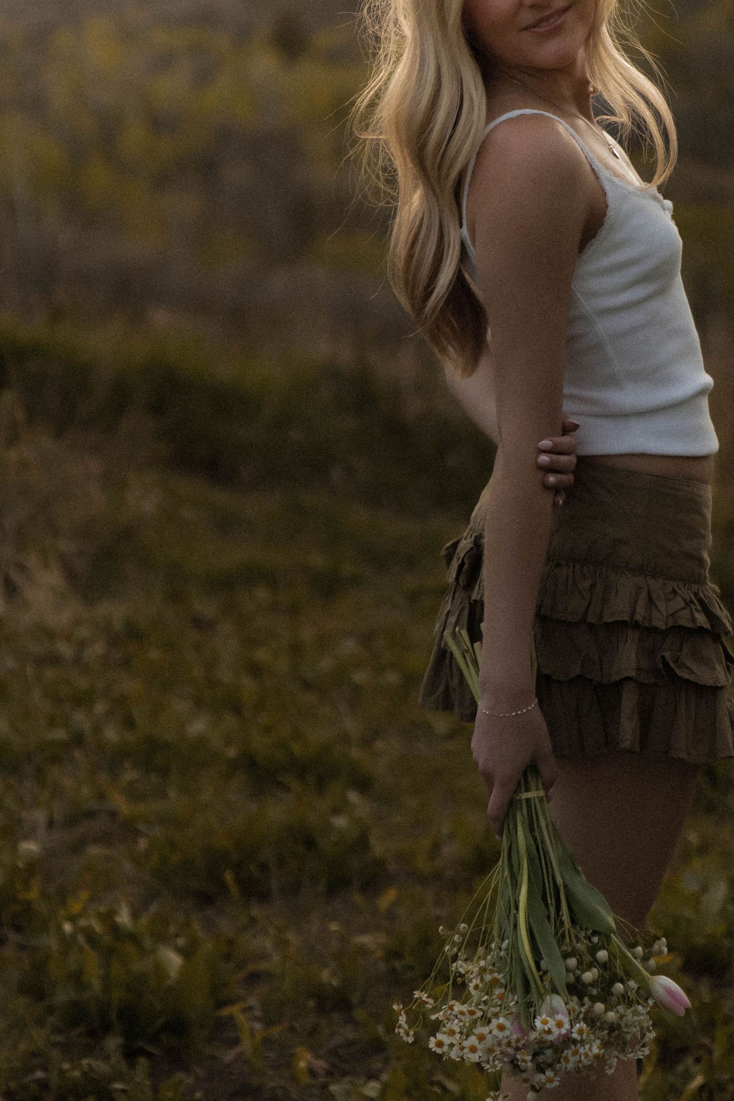 Blonde woman in white tank top and olive ruffle mini skirt holds wildflower bouquet in field at dusk.