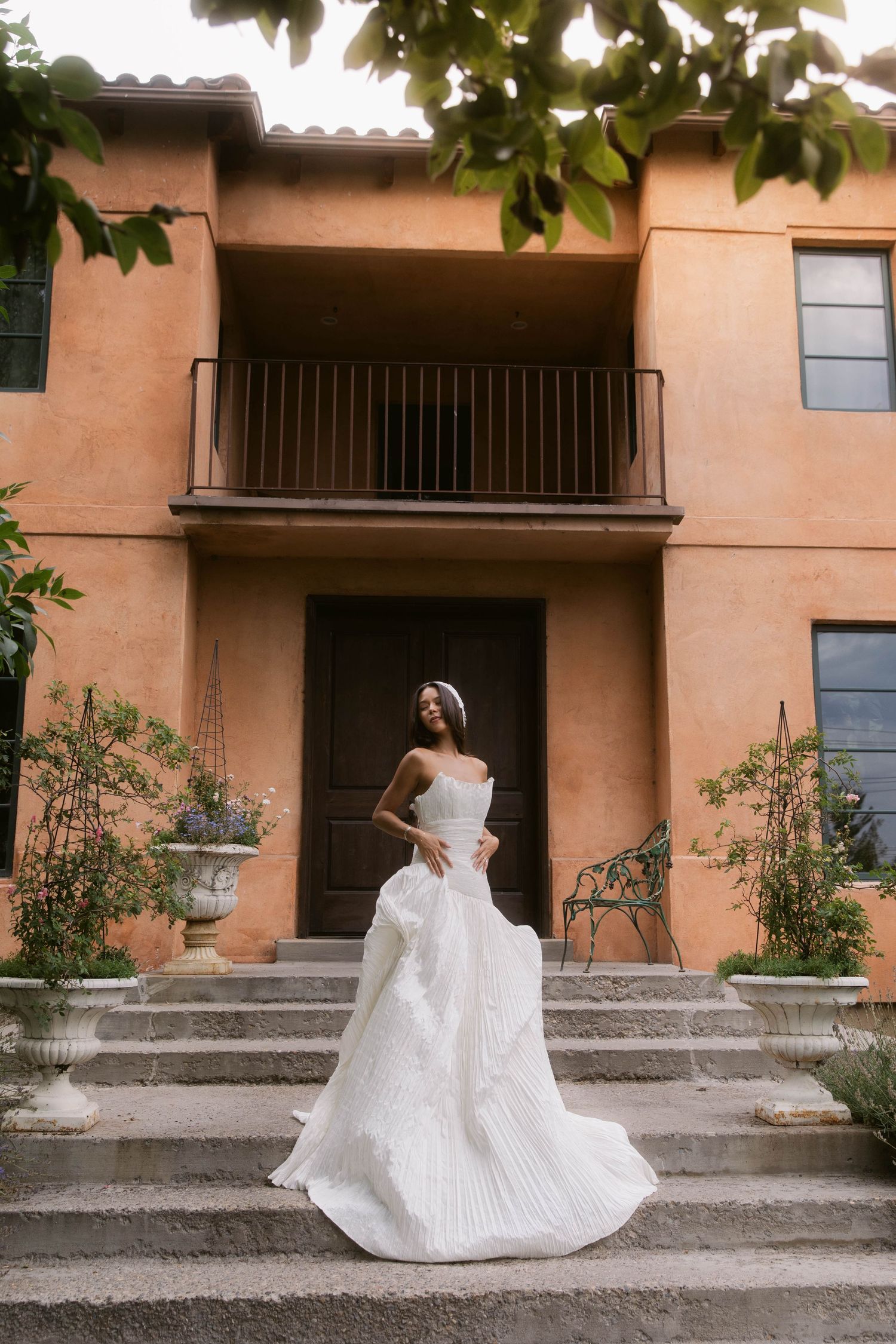 Bride in a strapless white pleated ballgown standing on steps outside a terracotta villa.