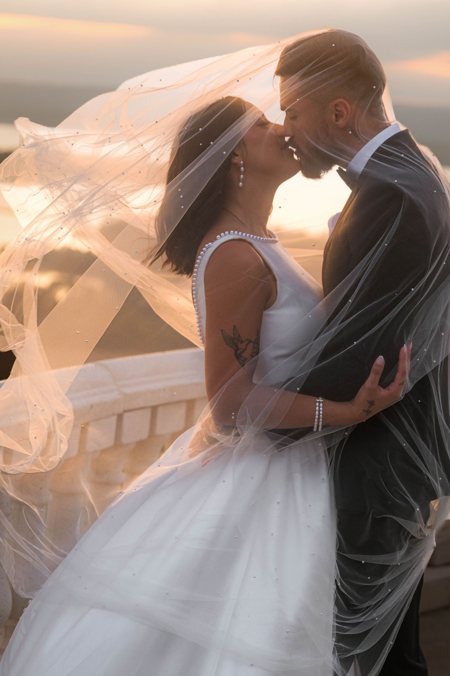 Bride and groom sharing a romantic kiss at sunset, with a flowing white veil billowing around them.