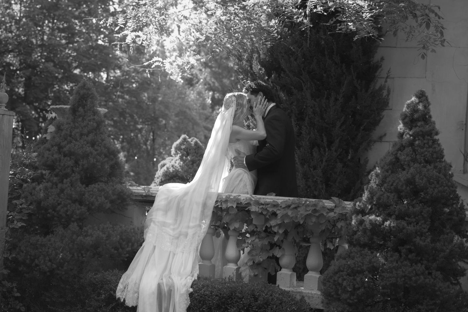 Black and white photo of bride and groom kissing on a stone balcony surrounded by garden greenery.