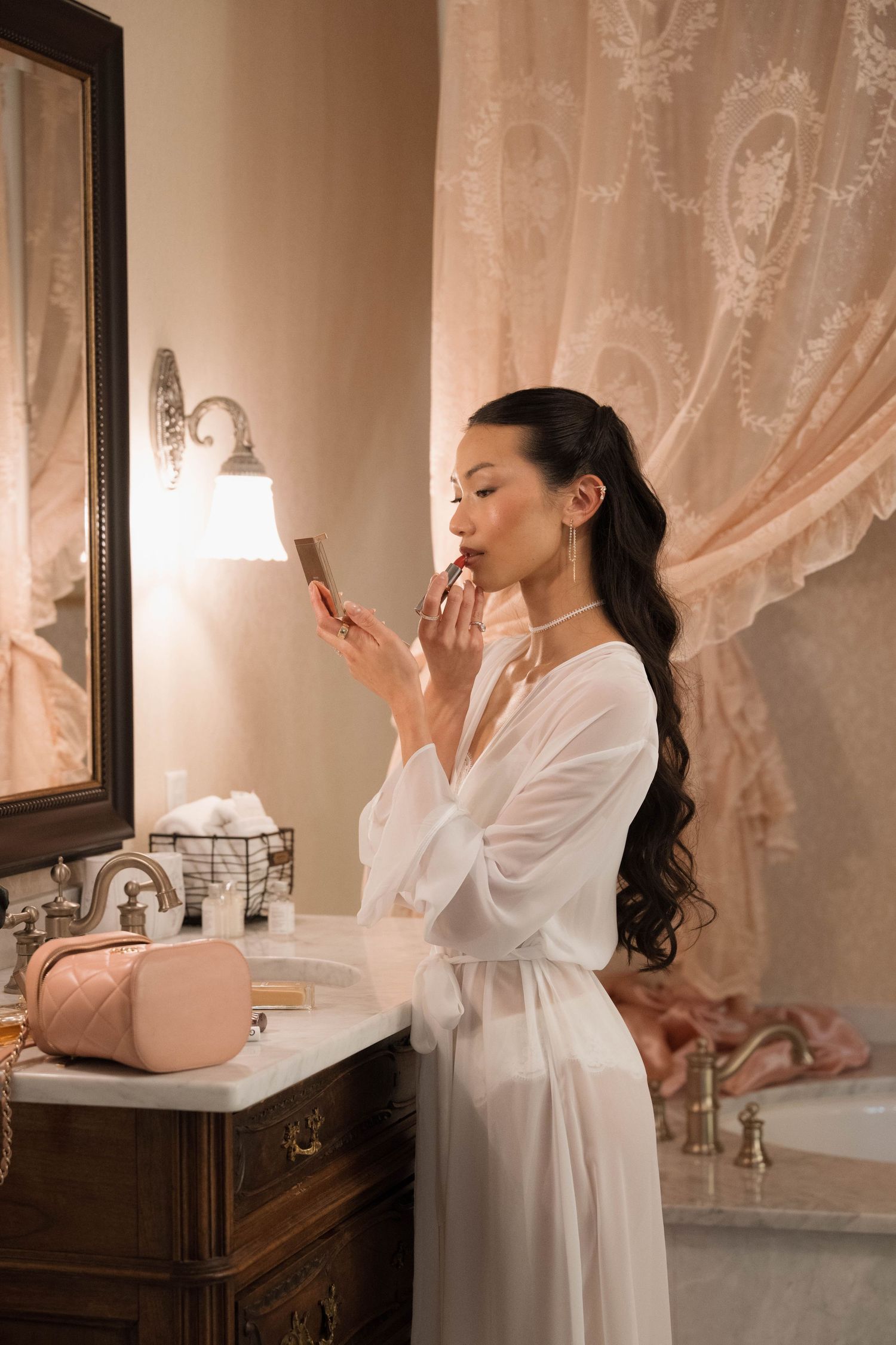 Bride in white robe applies lipstick using a hand mirror at a vintage bathroom vanity.