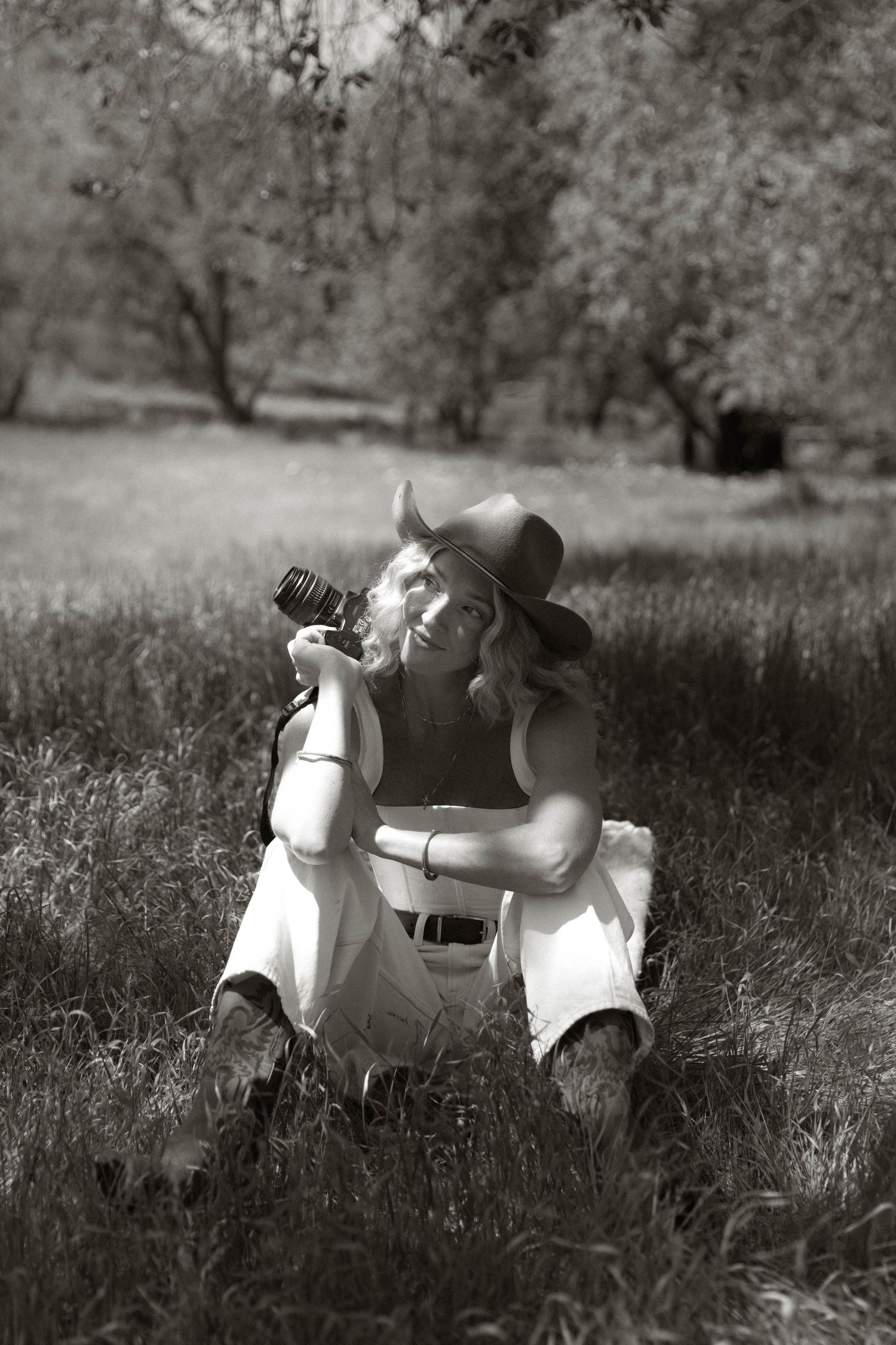 Black and white photo of a woman in a cowboy hat sitting in grass holding a vintage camera outdoors.