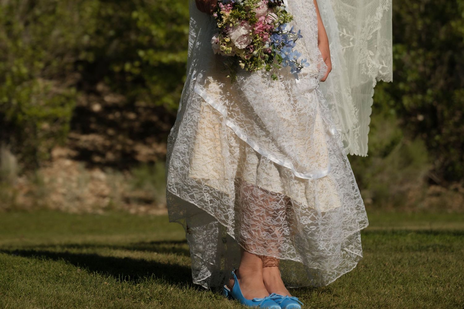 Close-up of bride in layered lace wedding dress wearing blue flats holding colorful wildflower bouquet.