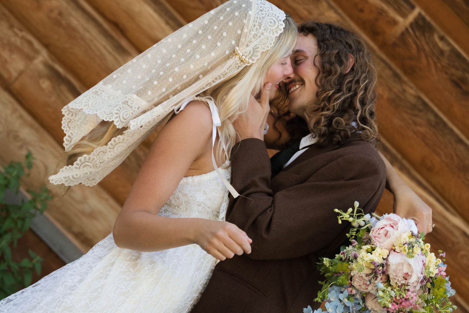 Bride and groom sharing a romantic moment, bride in lace dress with veil, groom in brown suit holding floral bouquet.